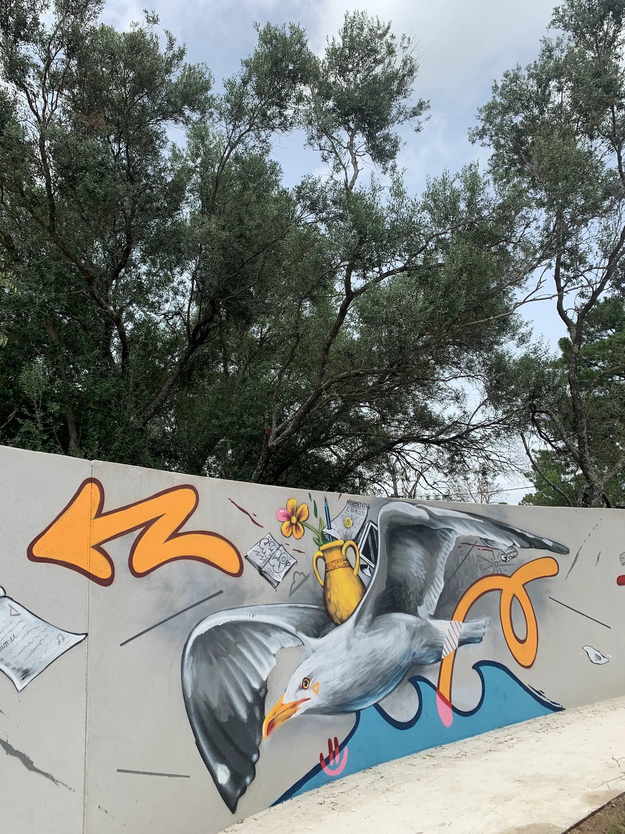 Fresque du jardin de la médiathèque L'animu, avec Piombu, 2021 : une mouette en vol, une amphore et une flèche orange peintes sur un mur courbe, sous les arbres