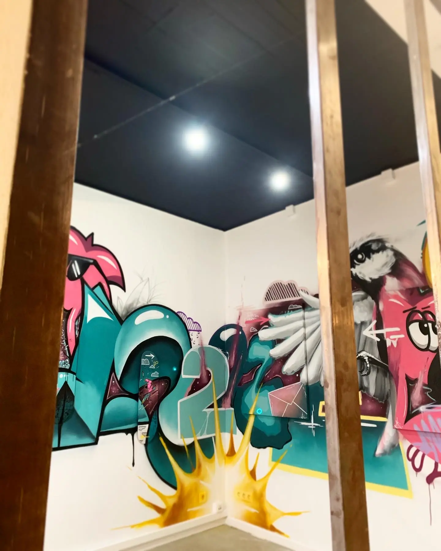 Exposition collective à La Conciergerie de Metz, 2020 : fresque graffiti couvrant deux murs d'angle, lettrage turquoise et personnages, vue à travers des montants en bois