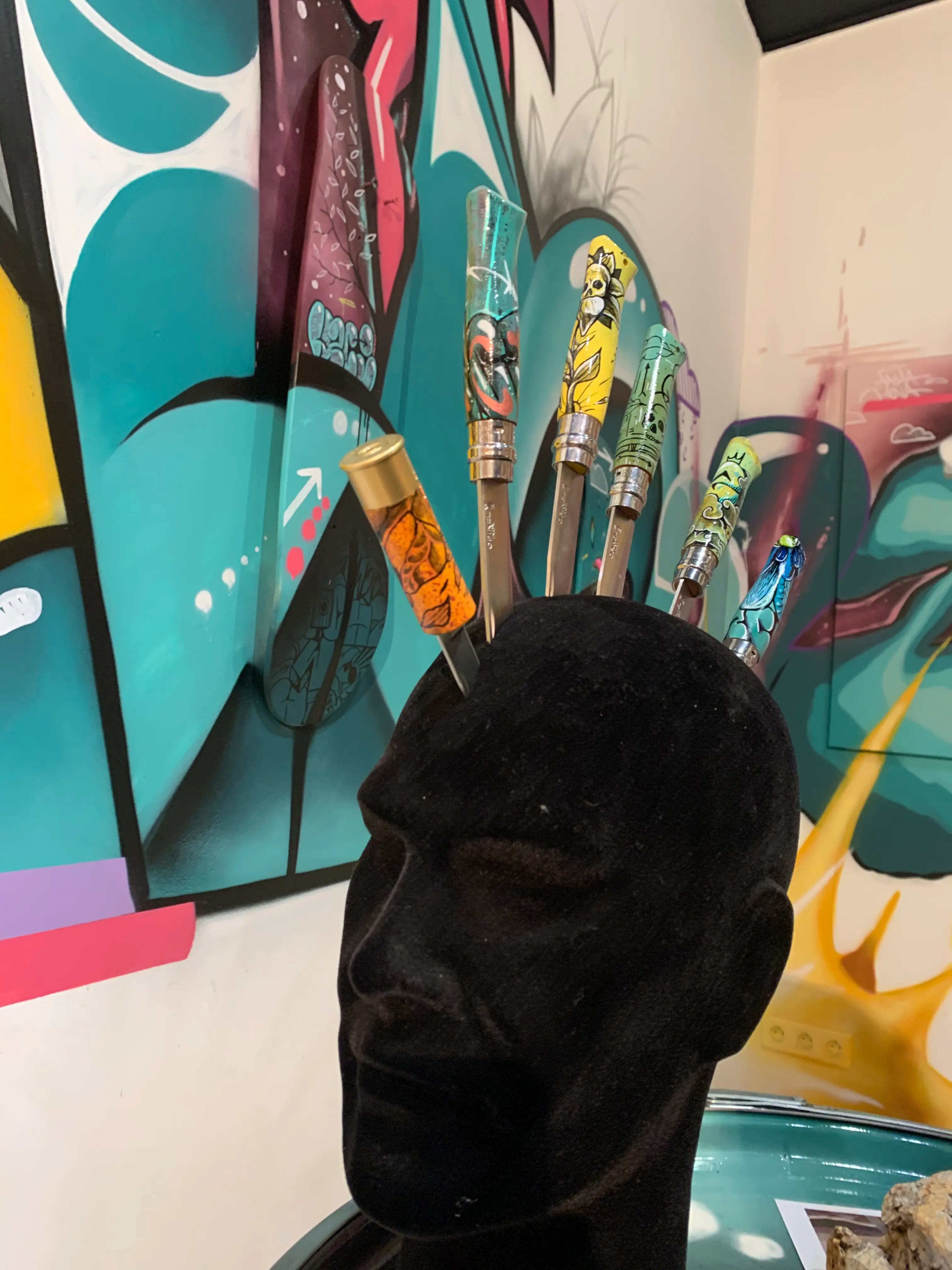 Exposition collective à La Conciergerie de Metz, 2020 : tête de mannequin noire hérissée de couteaux aux manches peints, devant la fresque turquoise