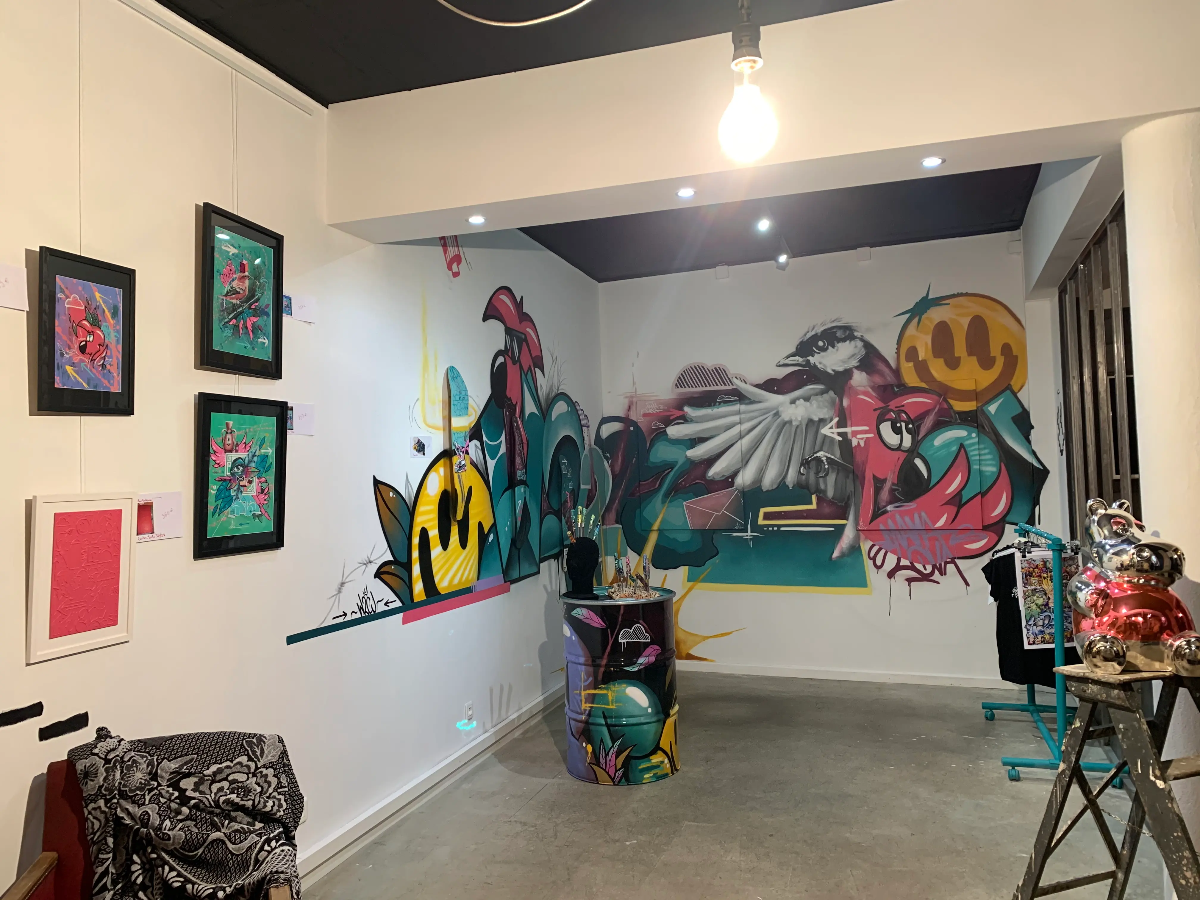 Exposition collective à La Conciergerie de Metz, 2020 : vue de la salle d'exposition, œuvres encadrées à gauche, fresque murale et bidon peint au fond