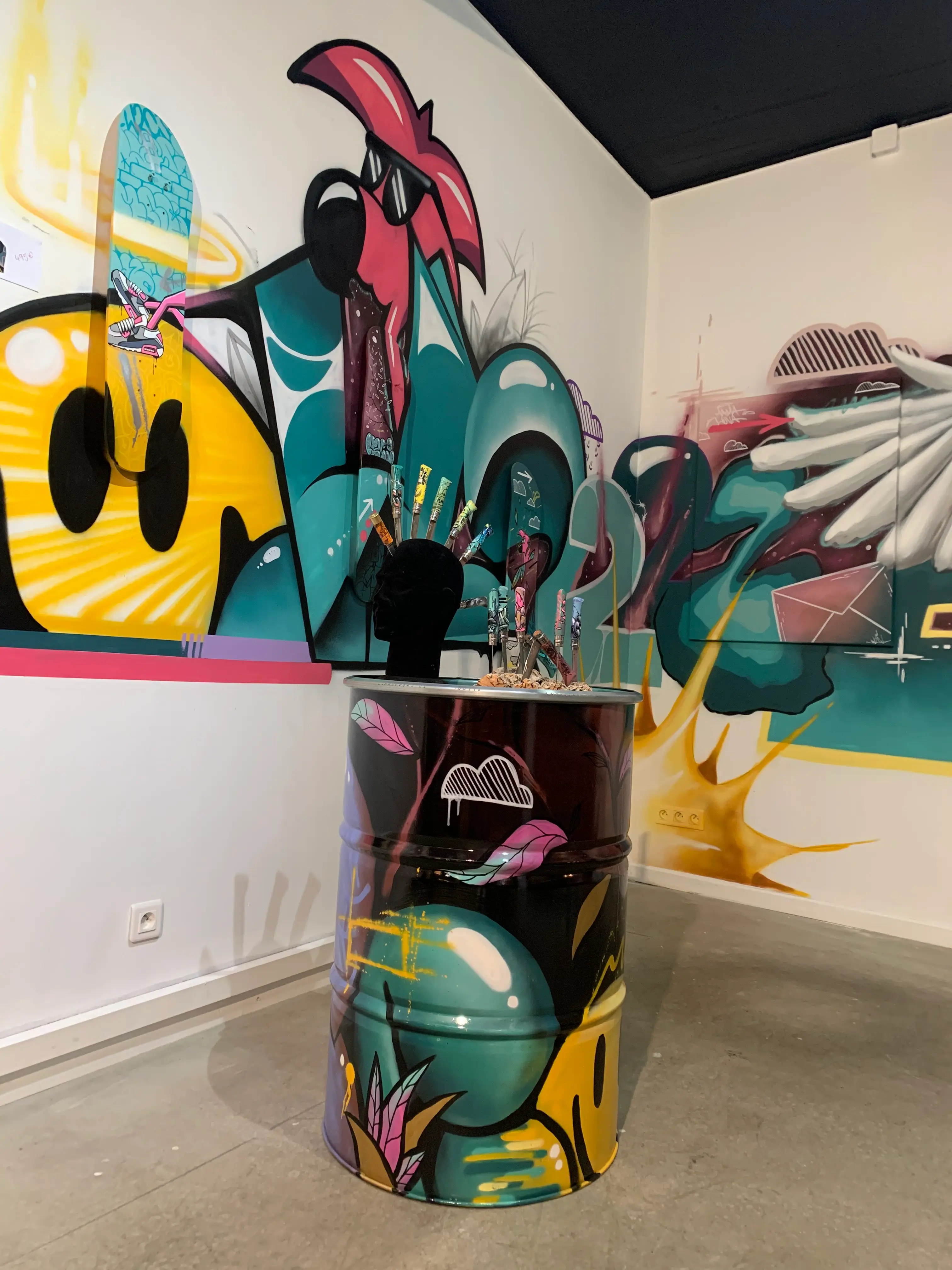 Exposition collective à La Conciergerie de Metz, 2020 : bidon peint surmonté d'une tête de mannequin et de couteaux peints, devant la fresque murale
