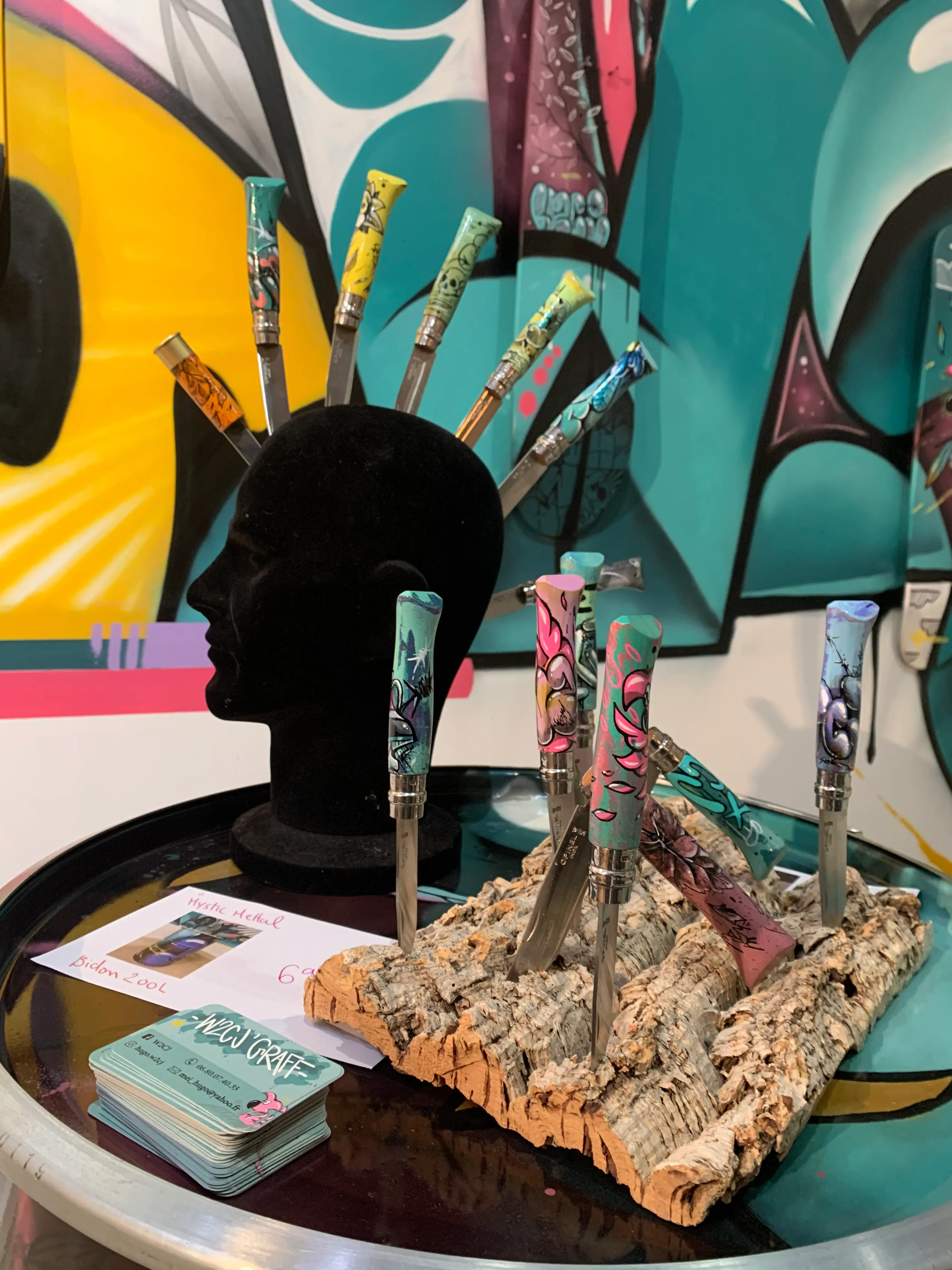 Exposition collective à La Conciergerie de Metz, 2020 : couteaux aux manches peints exposés sur un présentoir en liège et une tête de mannequin, devant la fresque