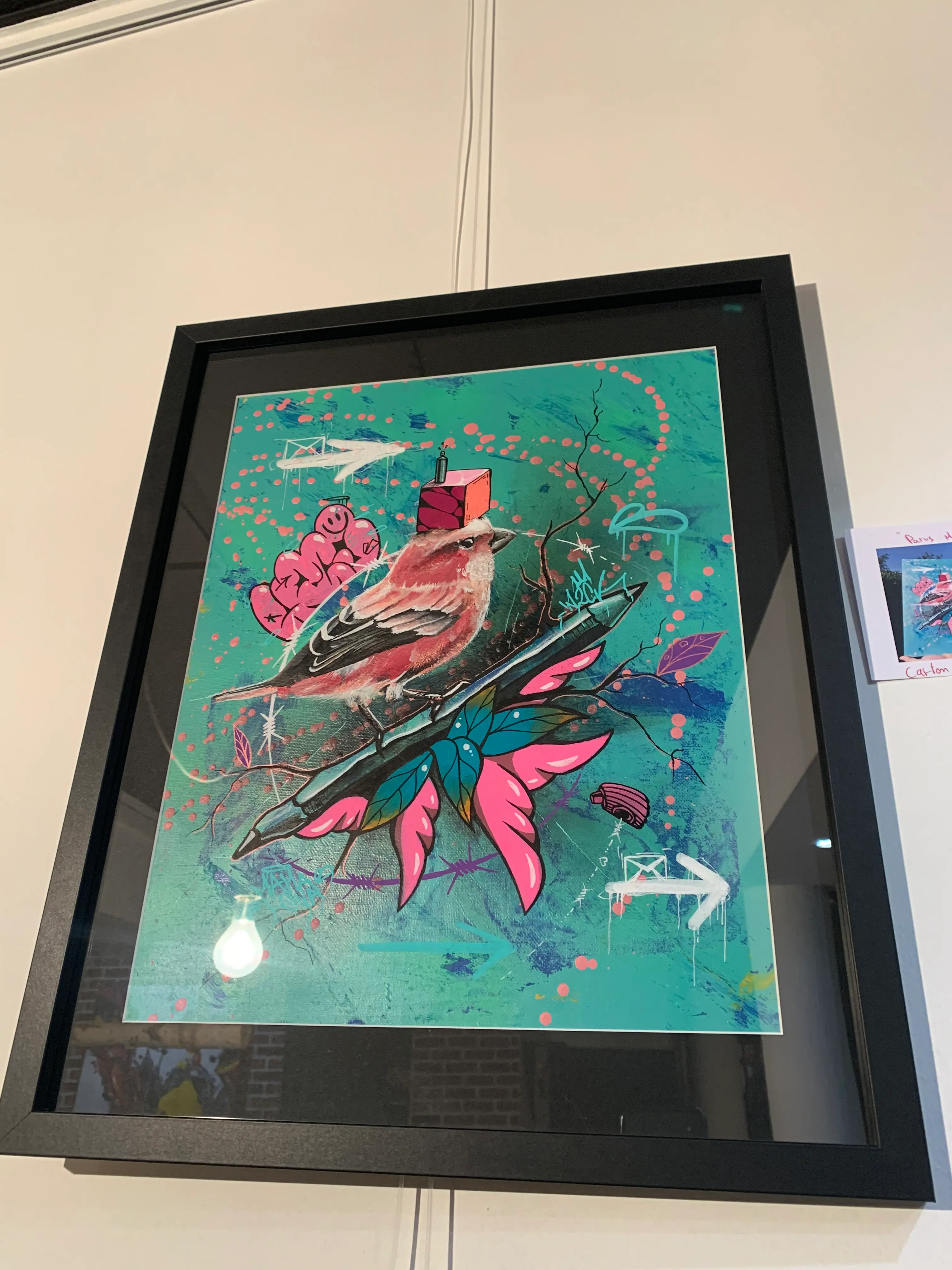 Exposition collective à La Conciergerie de Metz, 2020 : œuvre encadrée sur fond turquoise, un oiseau rose posé sur une bombe de peinture parmi des feuilles