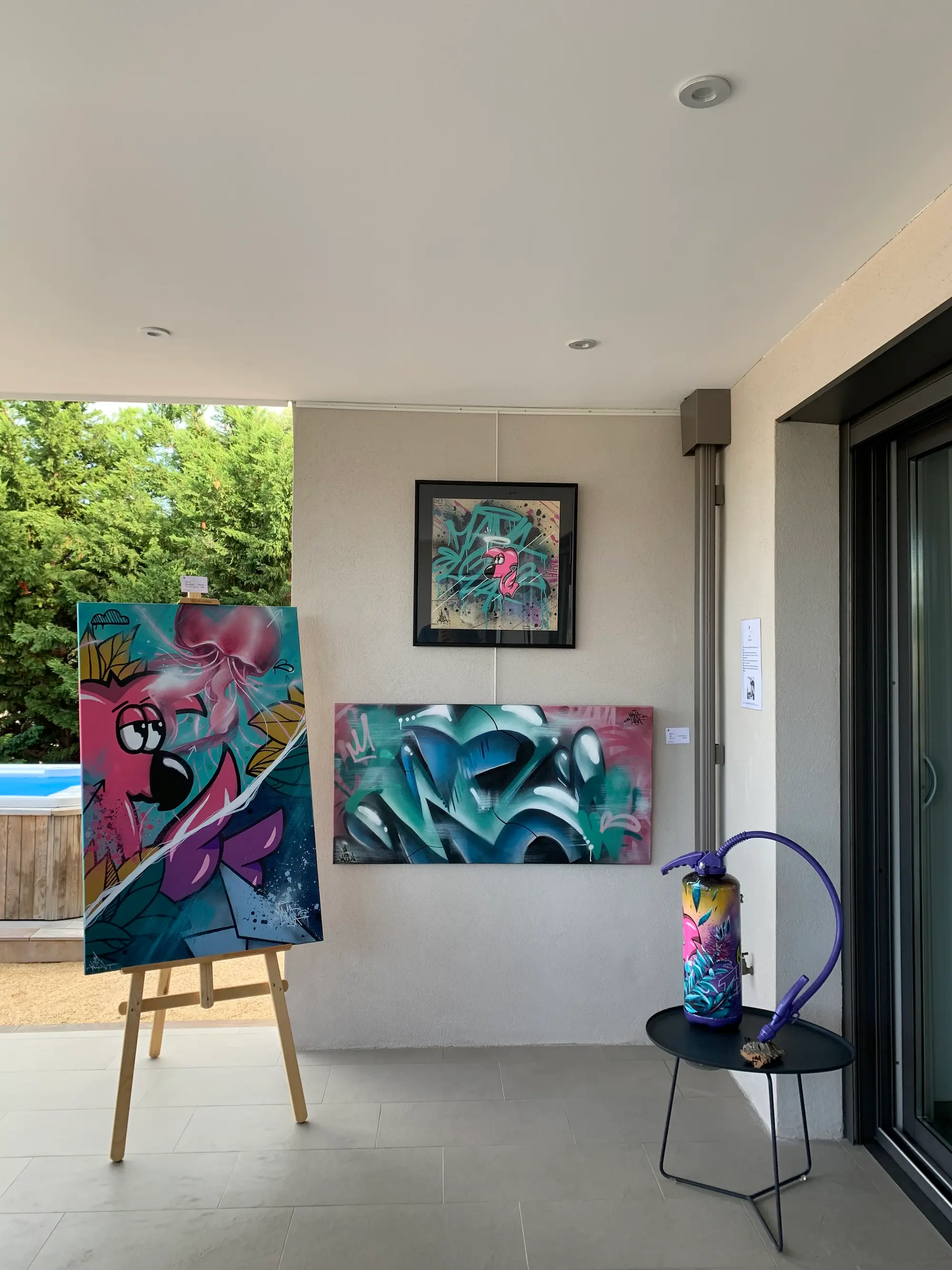 Résidence et exposition à la galerie Bel'arti, 2020 : accrochage sous la terrasse : toile de Roger le Flamant Rose sur chevalet, toile graffiti turquoise et extincteur peint