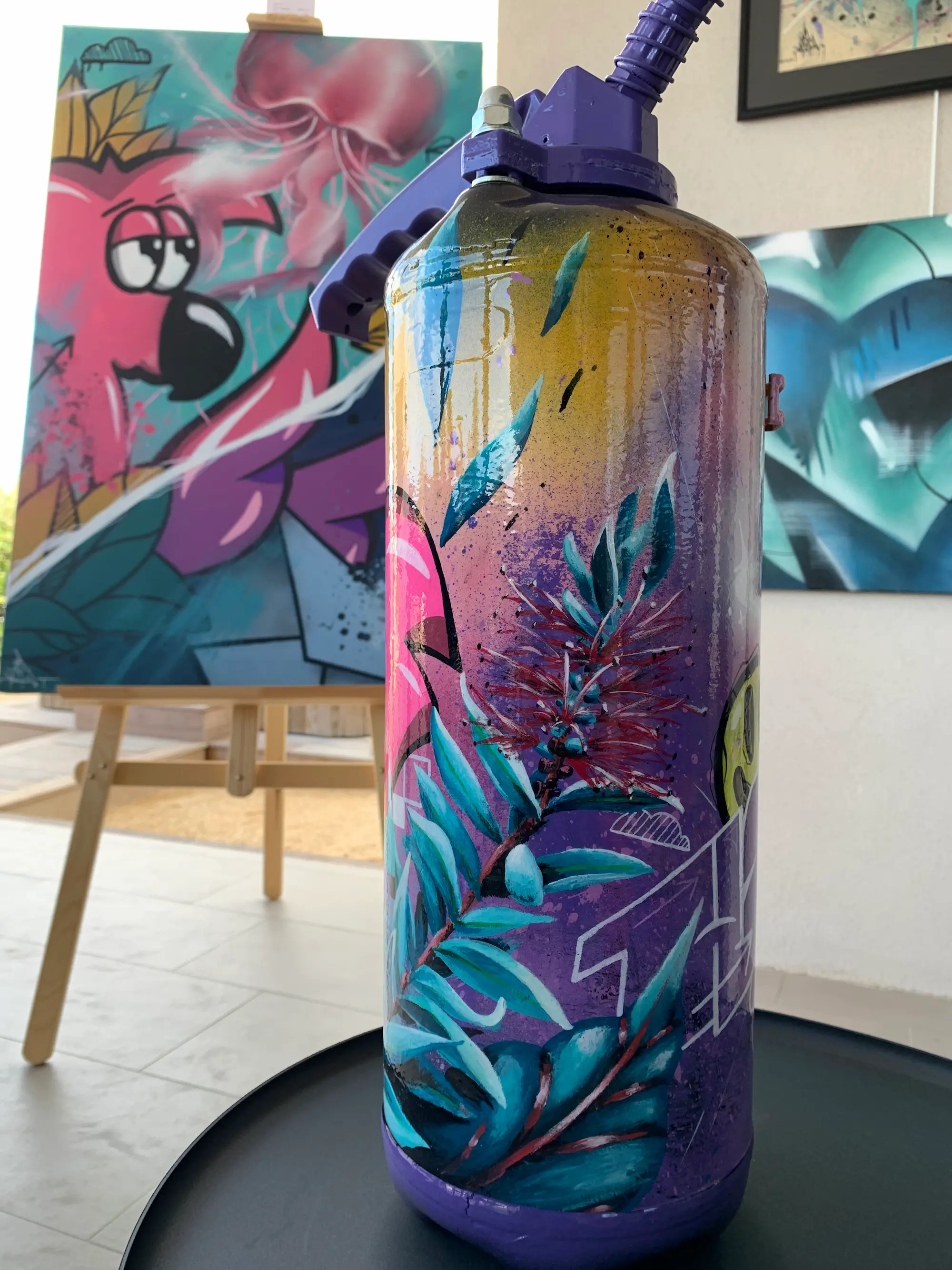 Résidence et exposition à la galerie Bel'arti, 2020 : extincteur peint en violet, motifs de feuillages turquoise, devant une toile de Roger le Flamant Rose sur chevalet