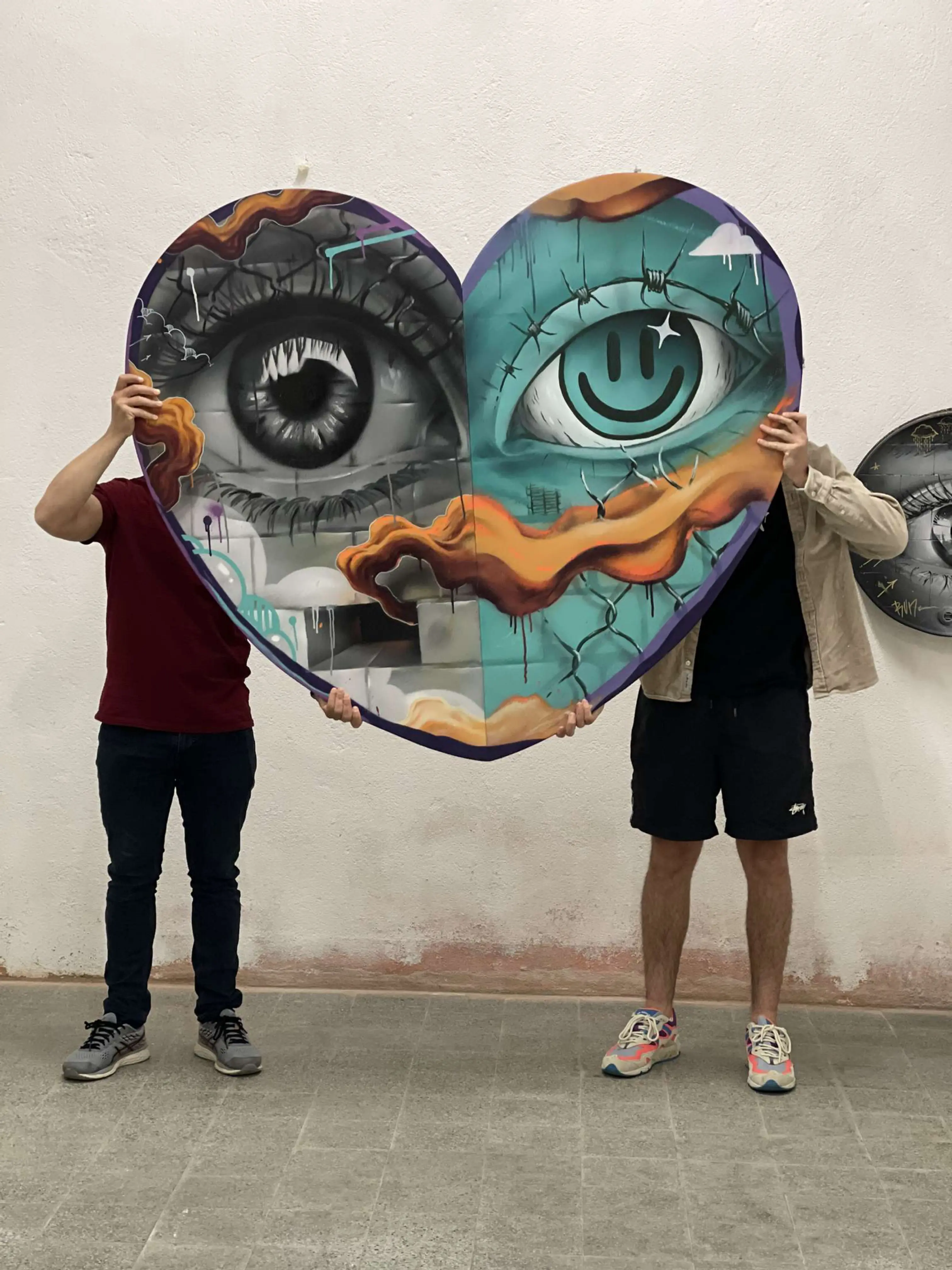Exposition en duo avec Karma Theora, galerie Noir & Blanc, 2021 : deux personnes tenant devant elles une grande toile en forme de cœur, un œil réaliste et un œil turquoise au regard smiley