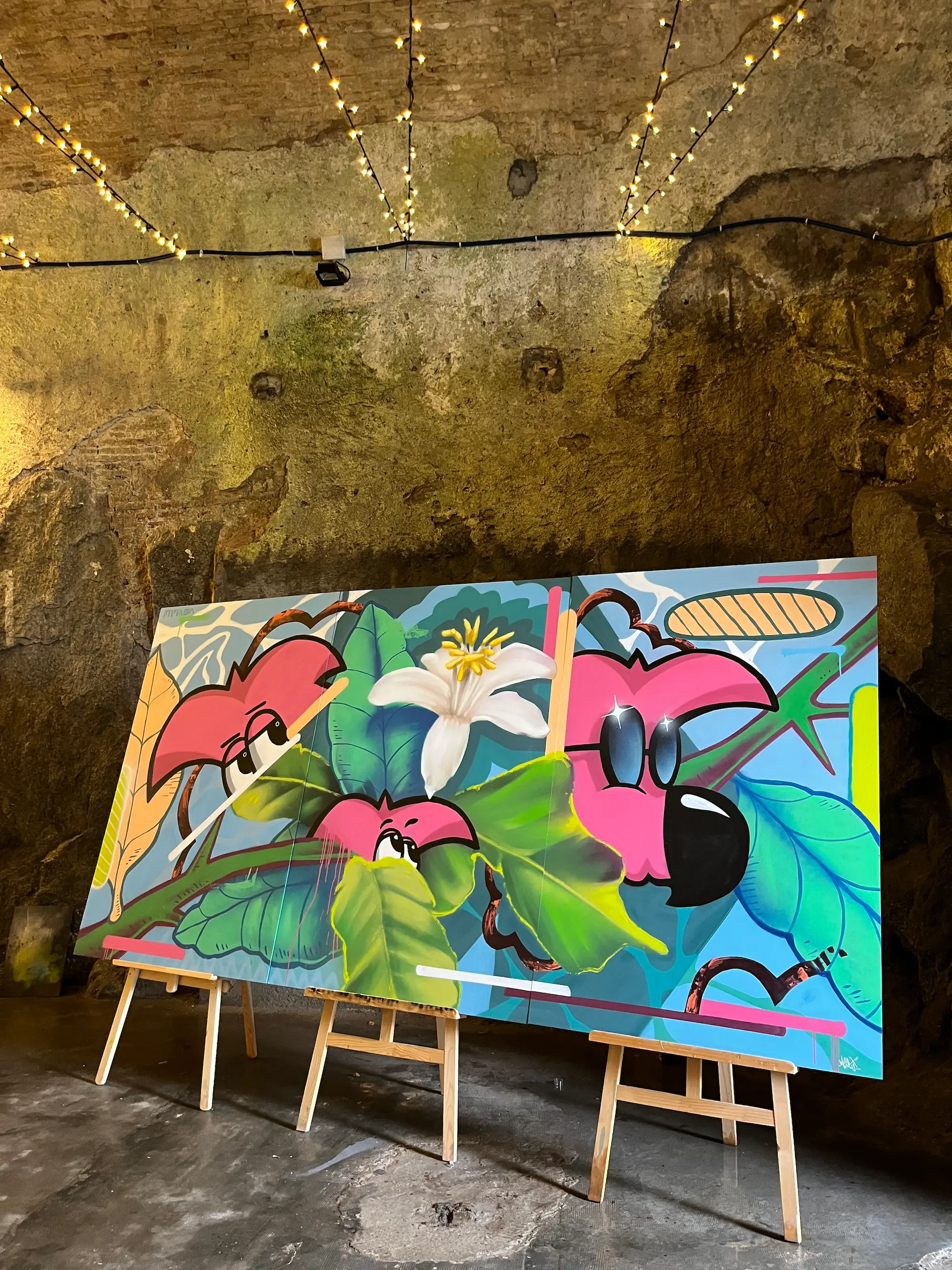 Live painting au festival Calviart, 2022 : grande toile sur chevalets dans une salle en pierre éclairée de guirlandes, Roger le Flamant Rose parmi les fleurs