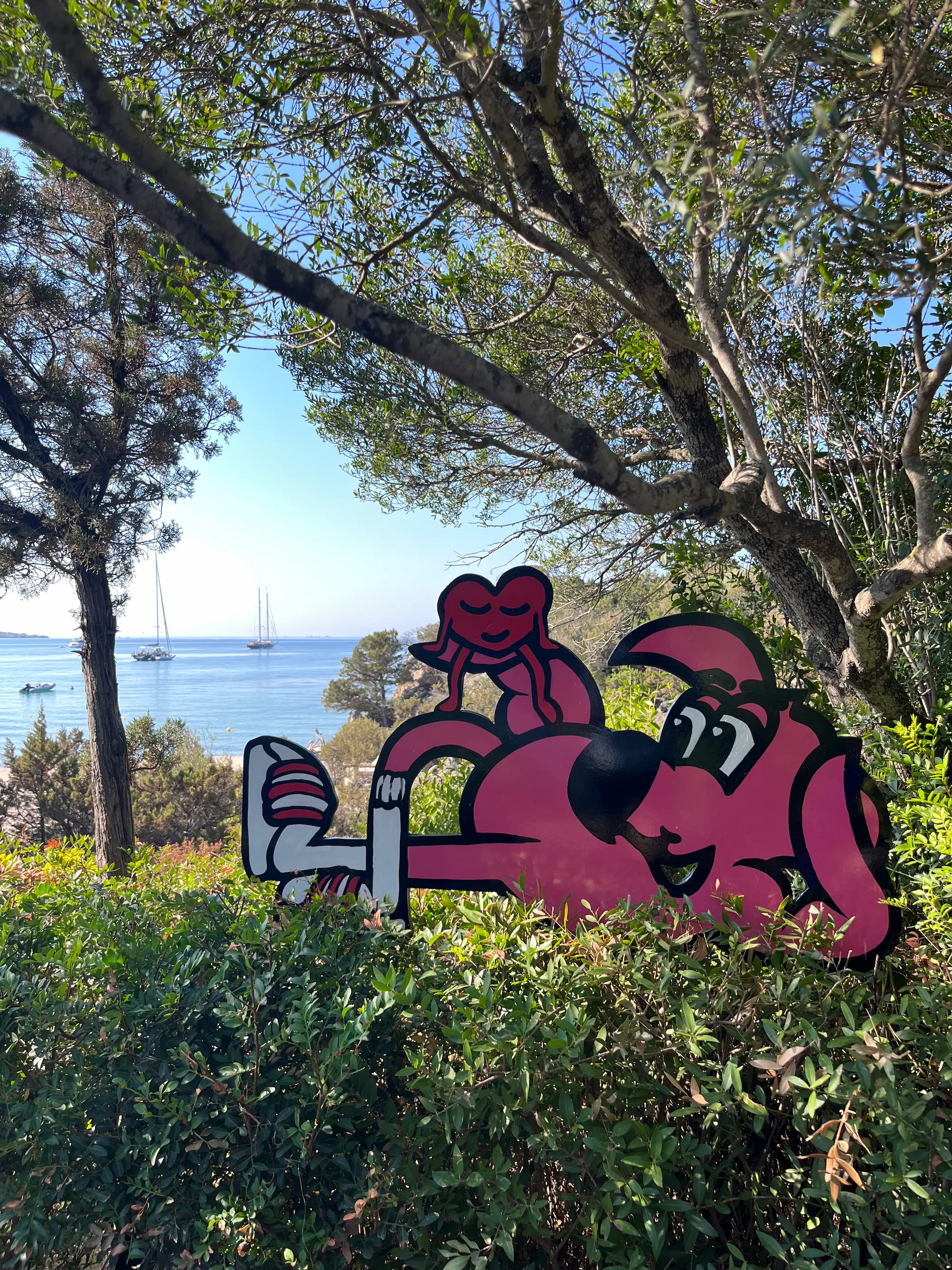 Exposition au Domaine de Murtoli, 2023 : découpe de Roger le Flamant Rose allongé dans la végétation, un cœur au-dessus de lui, la mer et des voiliers en arrière-plan