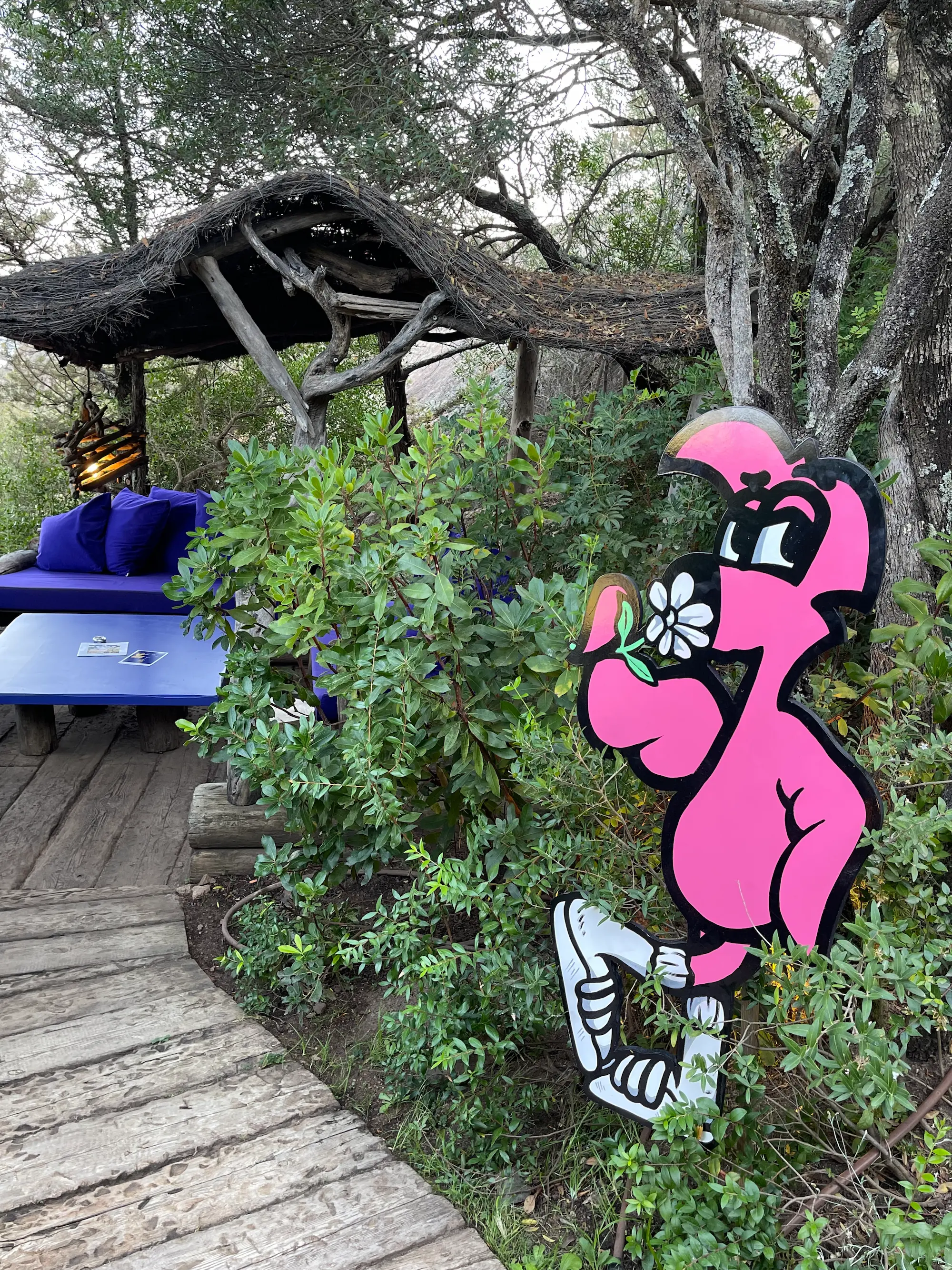 Exposition au Domaine de Murtoli, 2023 : découpe de Roger le Flamant Rose tenant une fleur, dans la végétation près d'une paillote