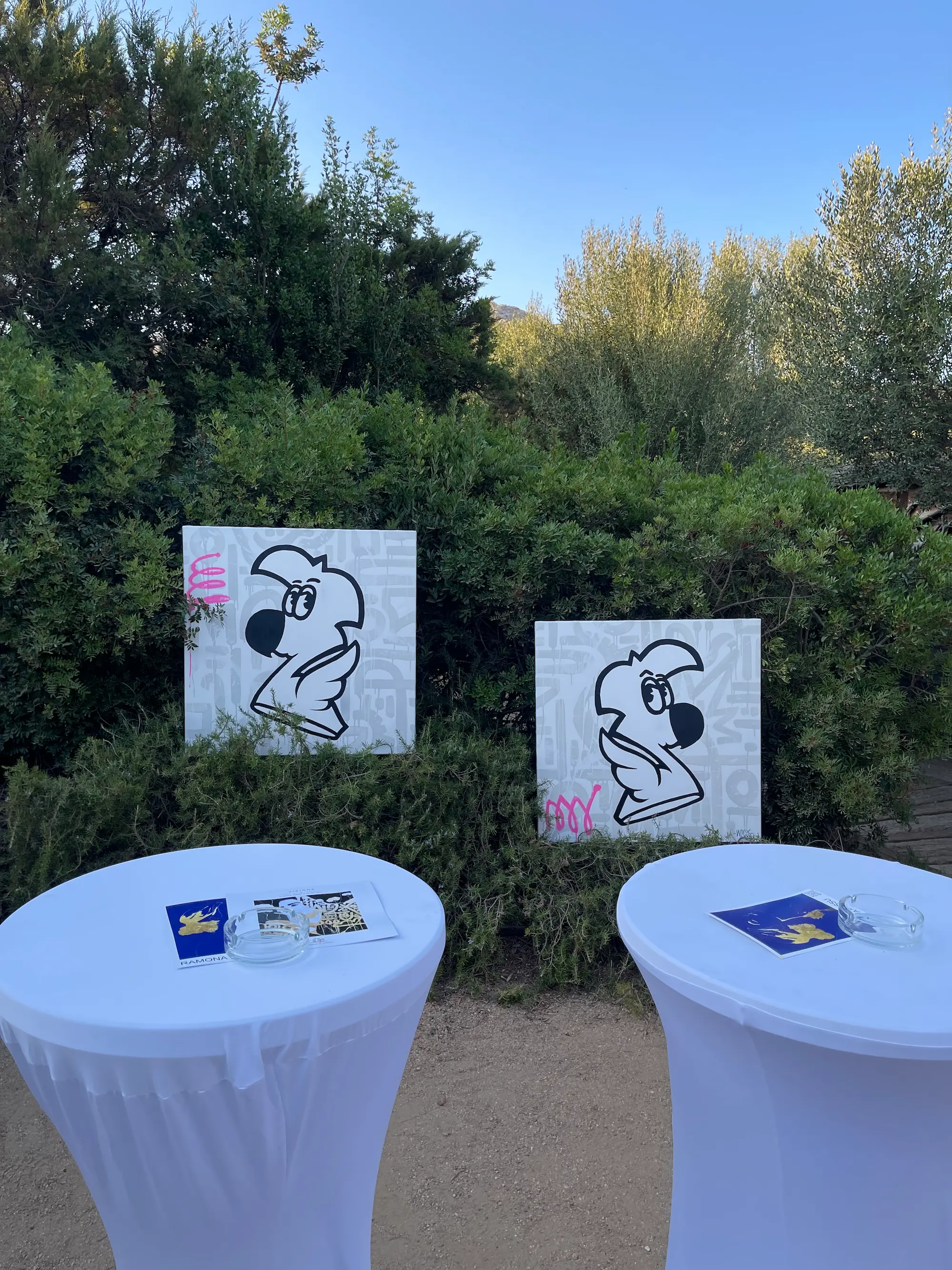 Exposition au Domaine de Murtoli, 2023 : deux toiles de Roger le Flamant Rose en noir et blanc posées dans la haie, tables hautes au premier plan