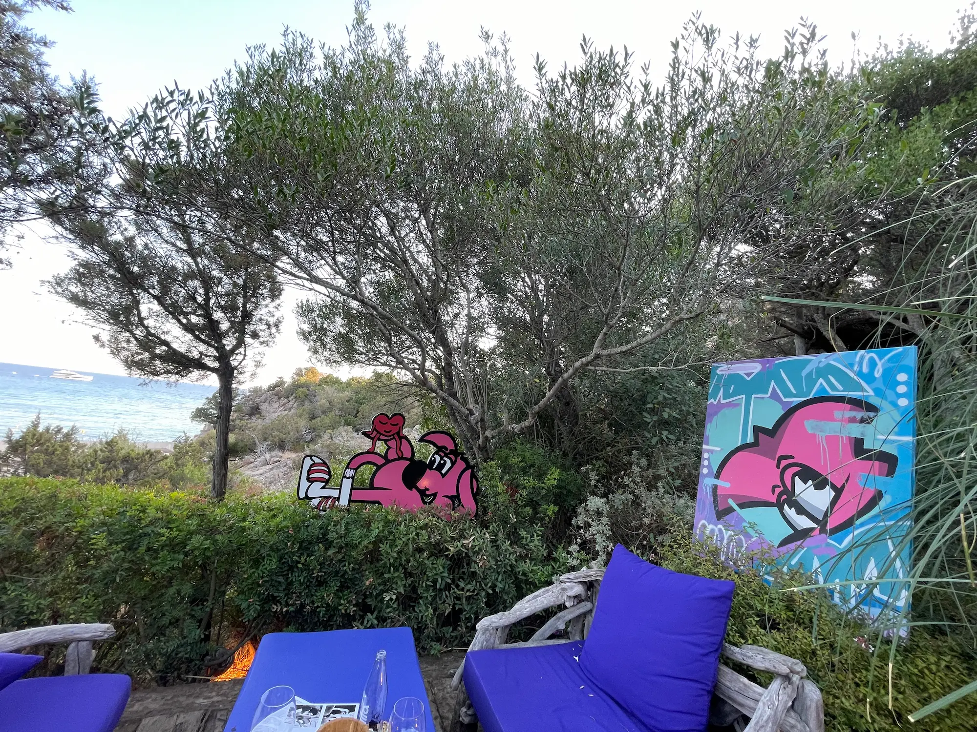 Exposition au Domaine de Murtoli, 2023 : découpe de Roger le Flamant Rose allongé et toile rose posée dans la végétation, coussins bleus au premier plan, la mer au loin