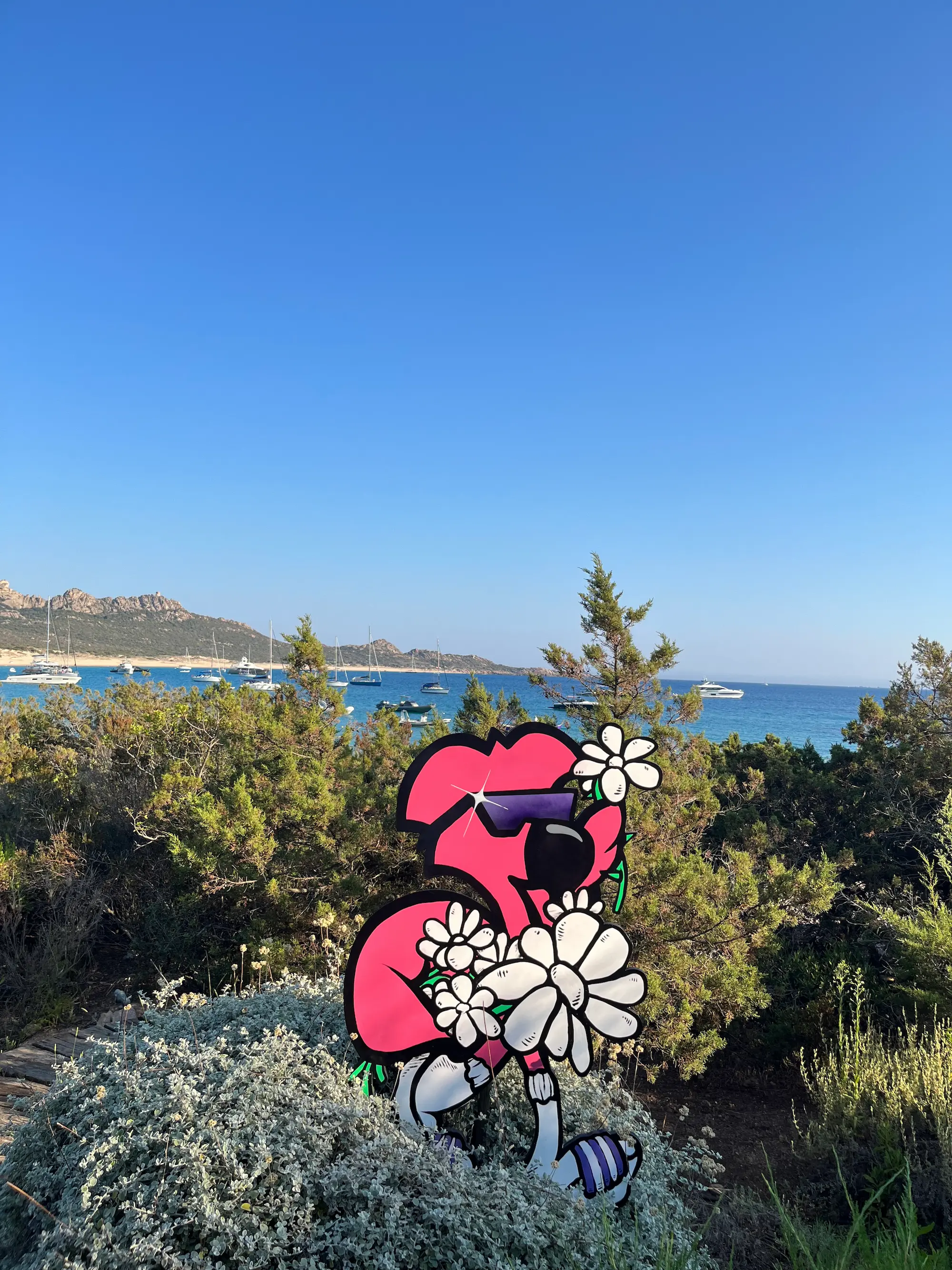 Exposition au Domaine de Murtoli, 2023 : découpe de Roger le Flamant Rose en lunettes, tenant des fleurs blanches, face à la baie et aux voiliers