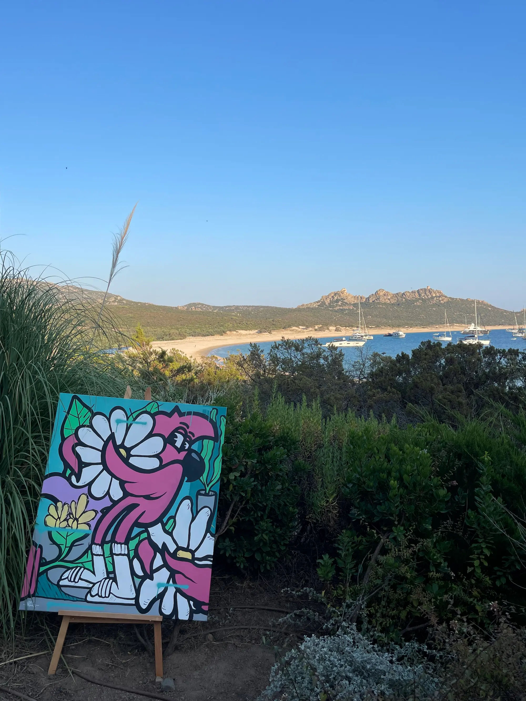 Exposition au Domaine de Murtoli, 2023 : toile de Roger le Flamant Rose parmi les fleurs sur un chevalet, face à la baie