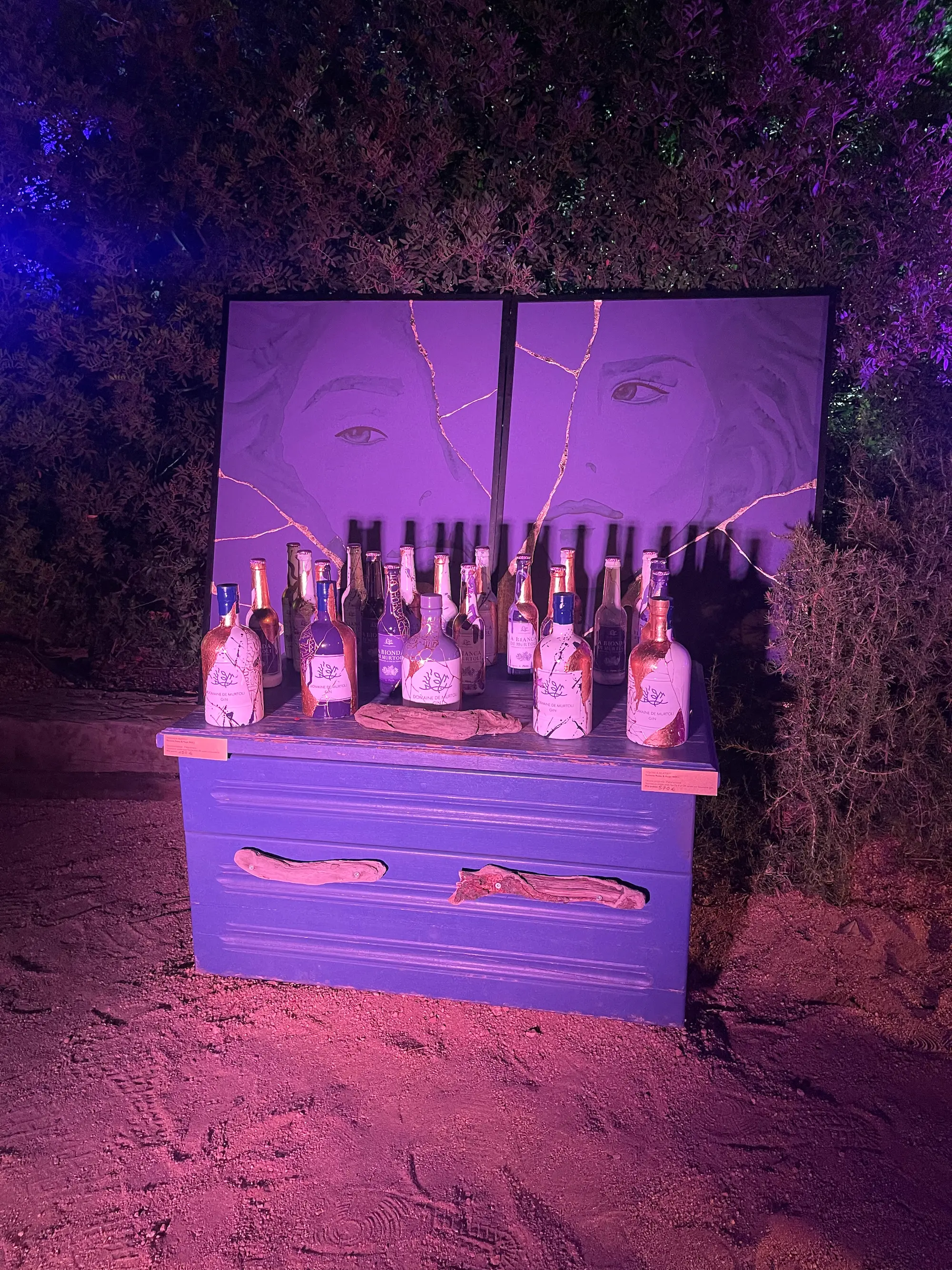 Exposition au Domaine de Murtoli, 2023 : commode peinte en violet chargée de bouteilles de gin, deux portraits sous éclairage violet, de nuit