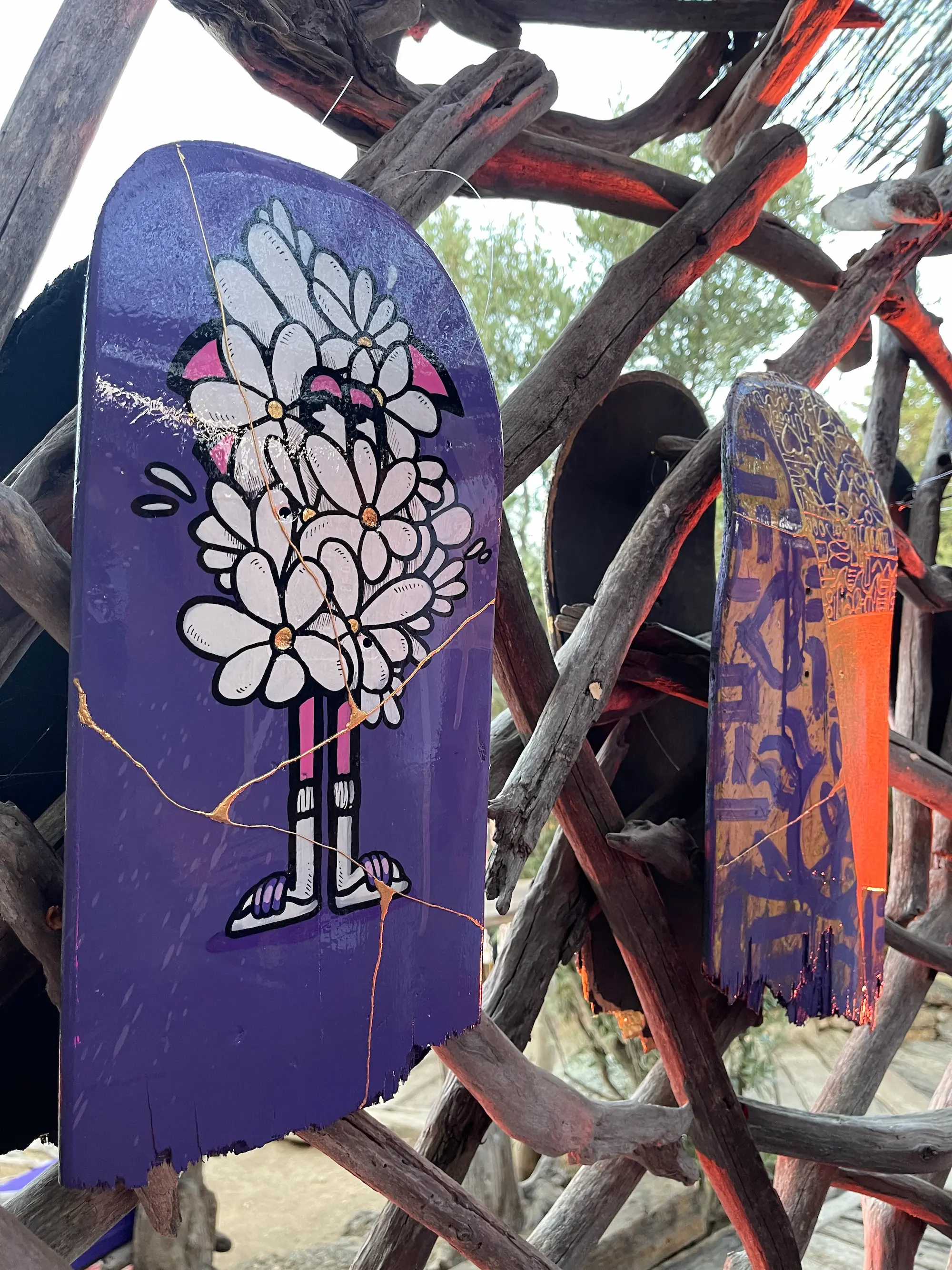 Exposition au Domaine de Murtoli, 2023 : plateaux de skate peints, un Roger le Flamant Rose caché derrière un bouquet sur fond violet, accrochés à des branches