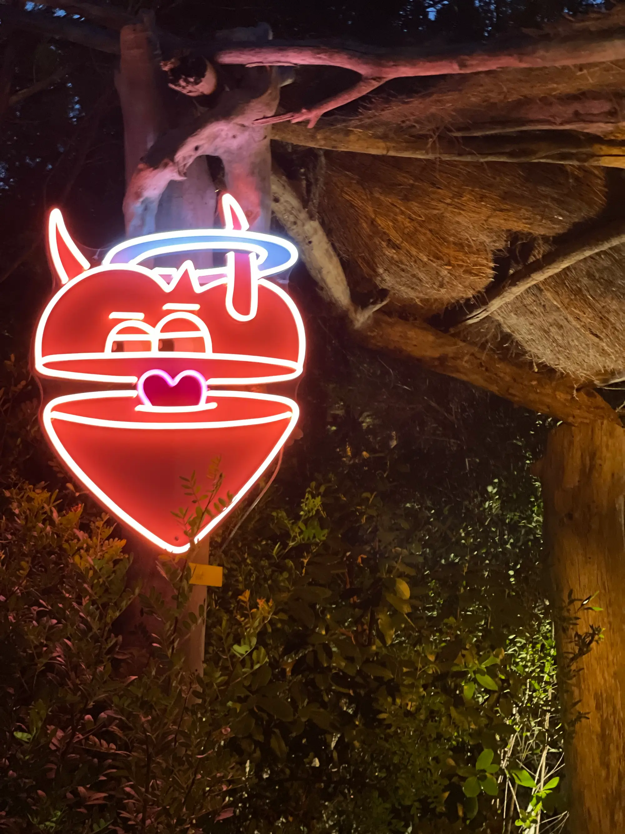 Exposition au Domaine de Murtoli, 2023 : néon en forme de cœur rouge mi-ange mi-démon, auréole et cornes, accroché sous un auvent de nuit