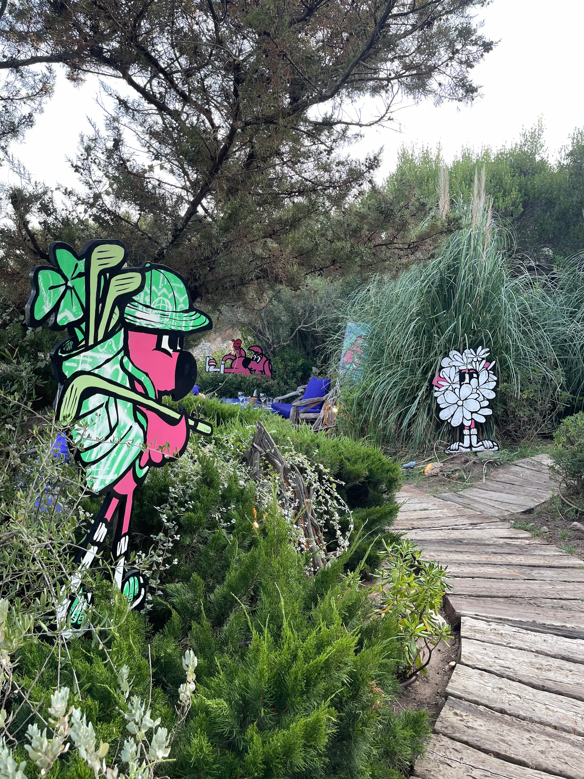 Exposition au Domaine de Murtoli, 2023 : découpe de Roger le Flamant Rose golfeur au bord d'un chemin en bois, autres découpes dans le maquis