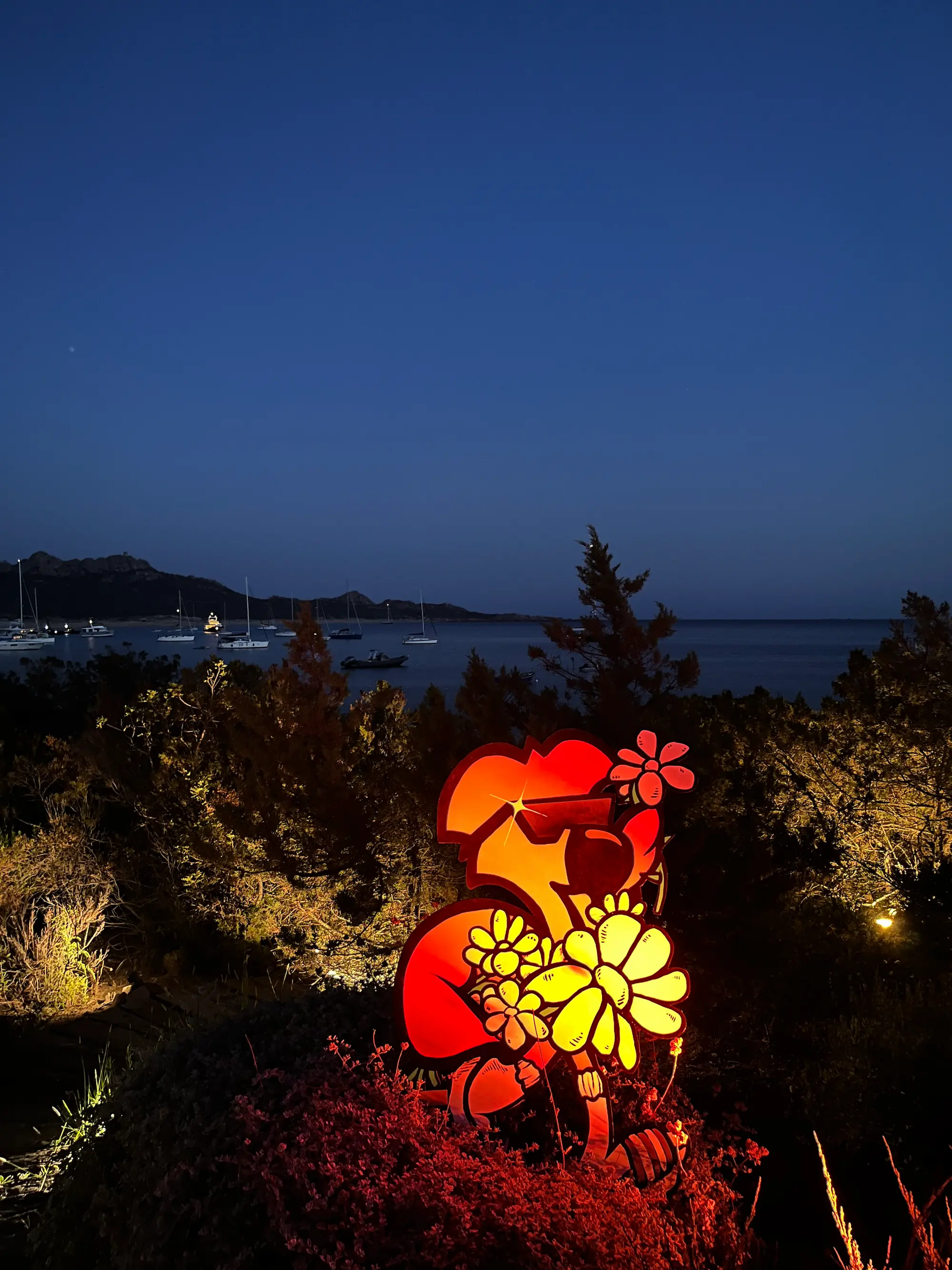Exposition au Domaine de Murtoli, 2023 : découpe lumineuse de Roger le Flamant Rose aux fleurs jaunes, face à la baie au crépuscule