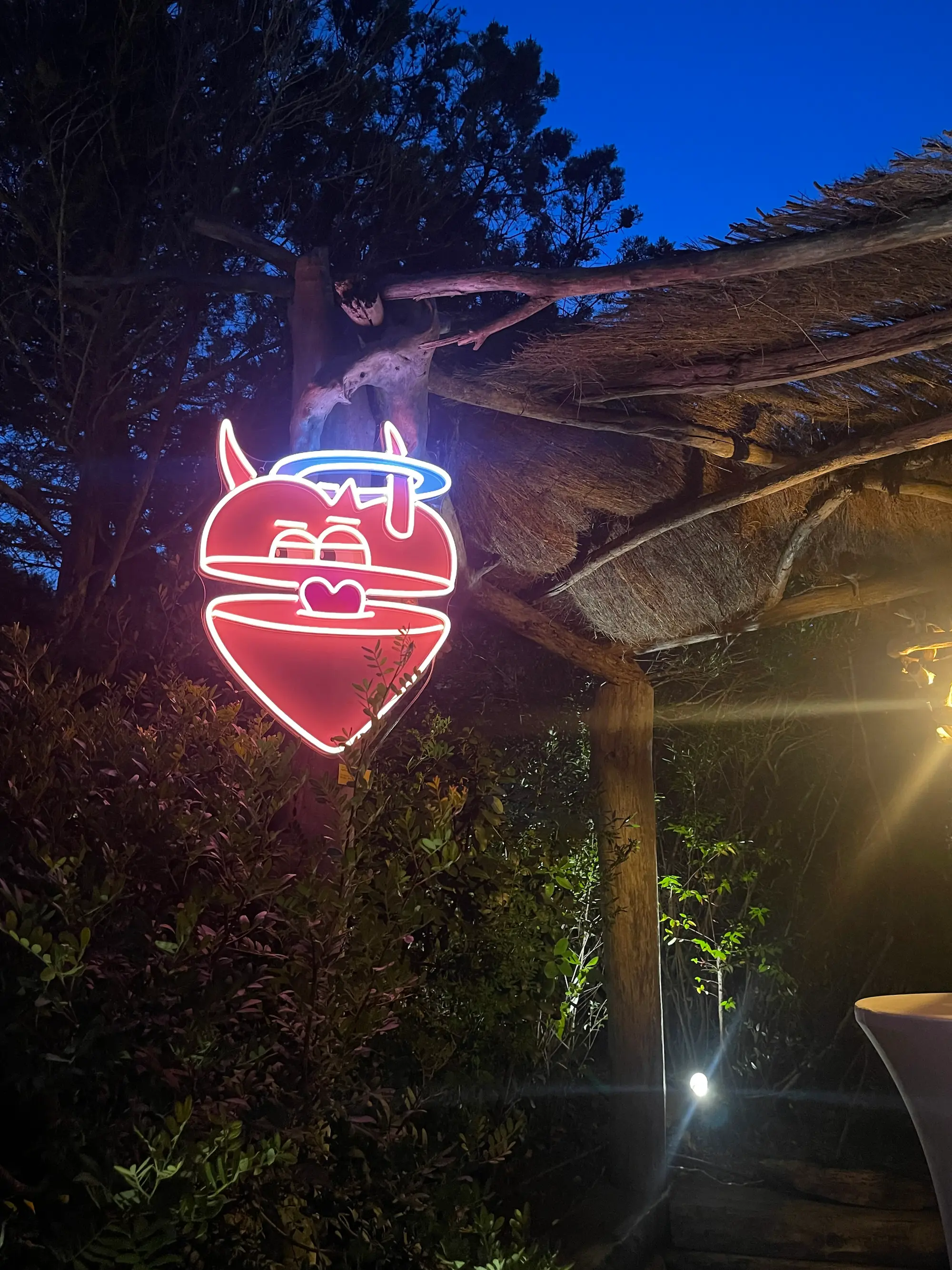 Exposition au Domaine de Murtoli, 2023 : néon du cœur mi-ange mi-démon allumé sous l'auvent de la paillote, à la tombée de la nuit