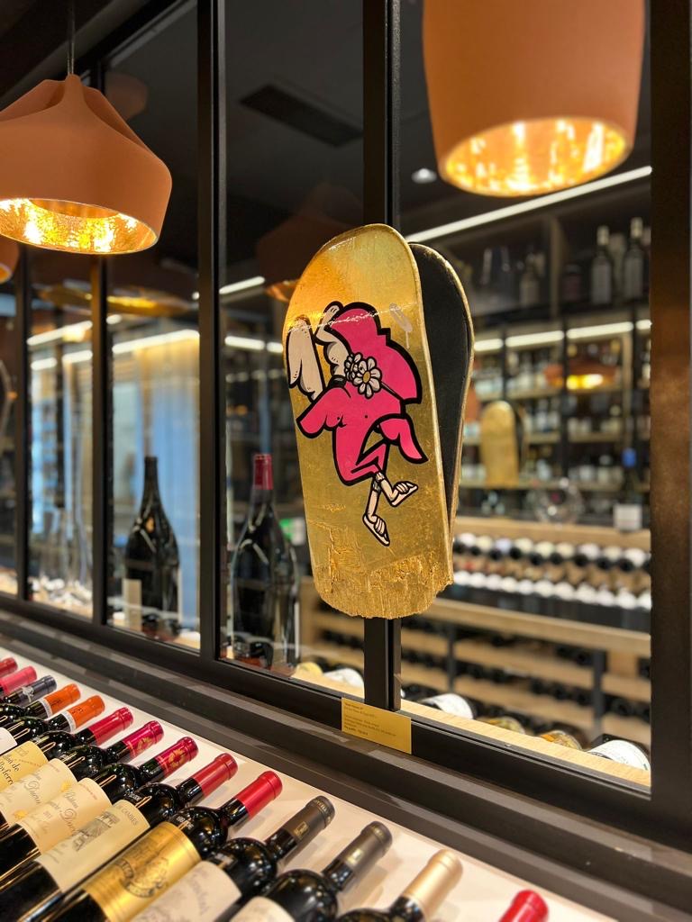 Exposition à L'Empreinte du vin, 2023 : plateau de skate doré peint d'un Roger le Flamant Rose, exposé dans une cave à vins