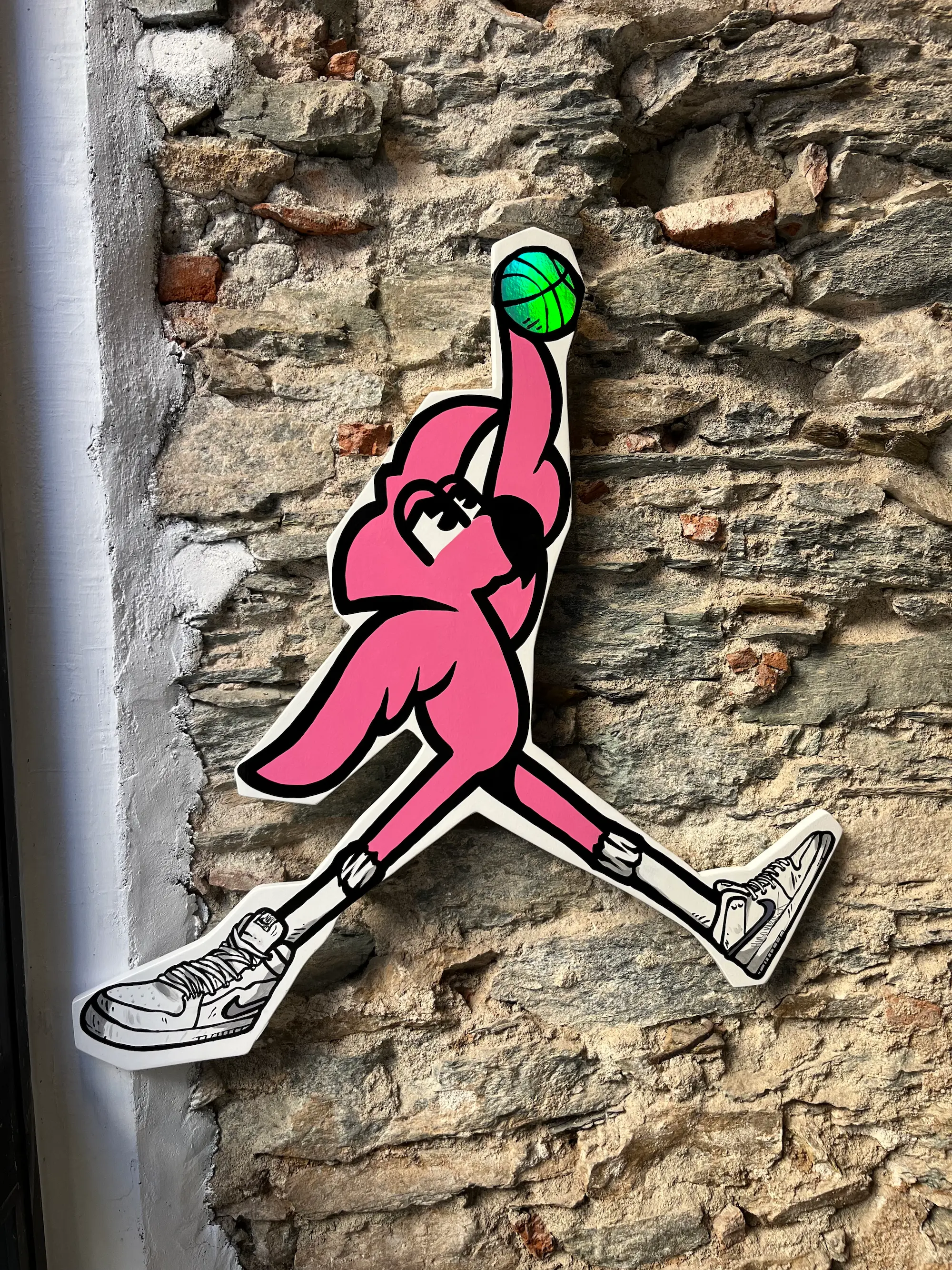 Exposition collective à la Capu Galerie, 2024 : découpe de Roger le Flamant Rose en basketteur, ballon vert à la main, sur un mur en pierre