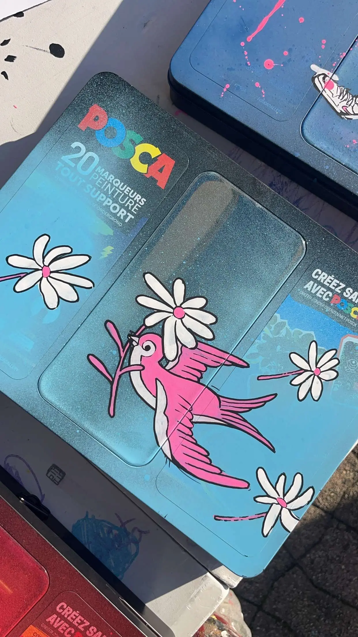 Collaboration Hugo W2CJ x Posca : coffret Posca bleu customisé, une hirondelle rose et des fleurs blanches