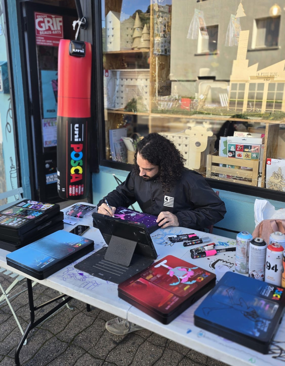Collaboration Hugo W2CJ x Posca : l'artiste customisant des coffrets Posca sur une table devant une papeterie