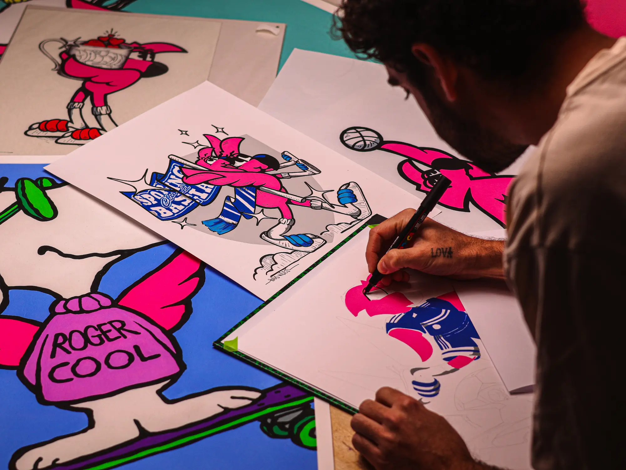 Collaboration Hugo W2CJ x Sporting Club de Bastia : l'artiste dessinant au feutre, dessins de Roger le Flamant Rose étalés sur la table