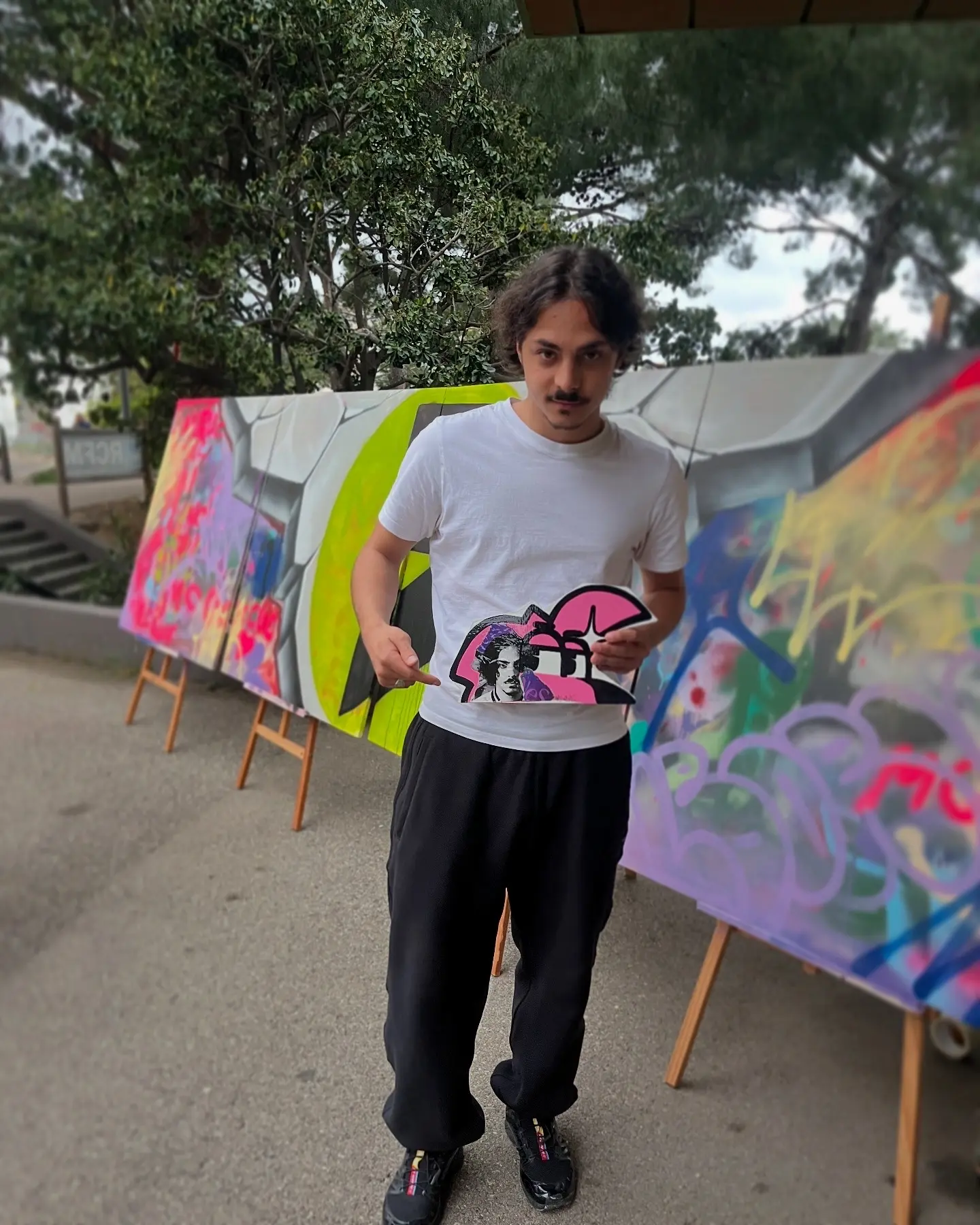 NeS, artiste du festival East Side, tenant sa sculpture murale de Roger le Flamant Rose devant les panneaux du live painting de Hugo W2CJ