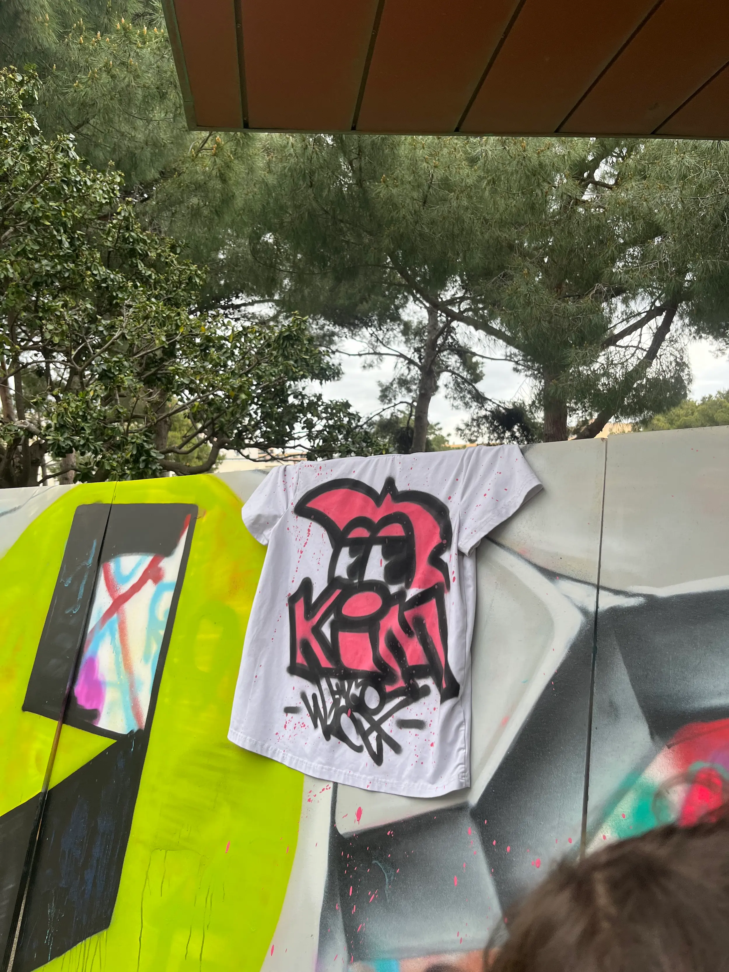 T shirt blanc graffé en rose et noir par Hugo W2CJ, suspendu devant le live painting du festival East Side