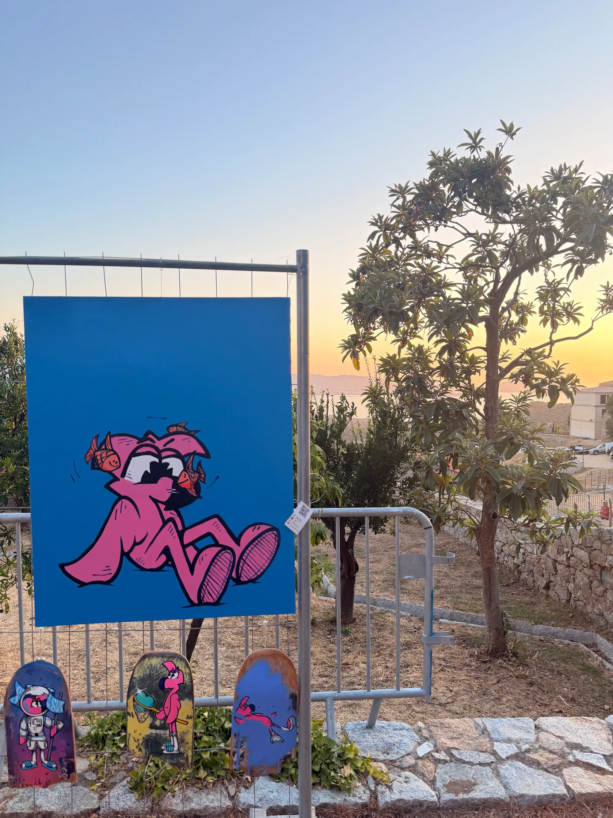 Toile bleue de Roger le Flamant Rose assis, accrochée en plein air au coucher du soleil, avec trois skates peints, festival Itinérances à Lumio