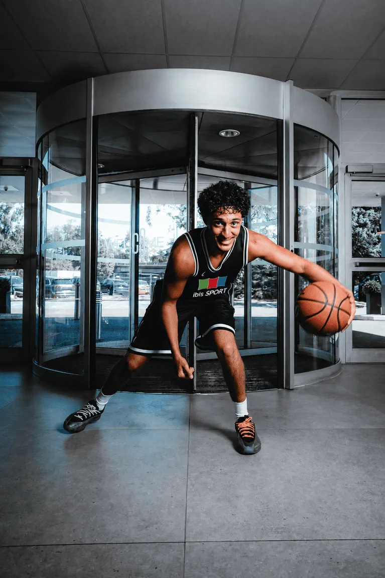 Un jeune homme en tenue de basketball noire dribble un ballon de basket devant une porte tournante en verre dans un espace intérieur lumineux.
