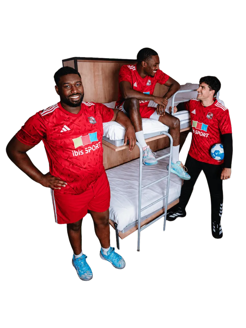 Trois hommes en tenues sportives rouges discutent près d'un lit superposé, l'un tenant un ballon de handball bleu et blanc.