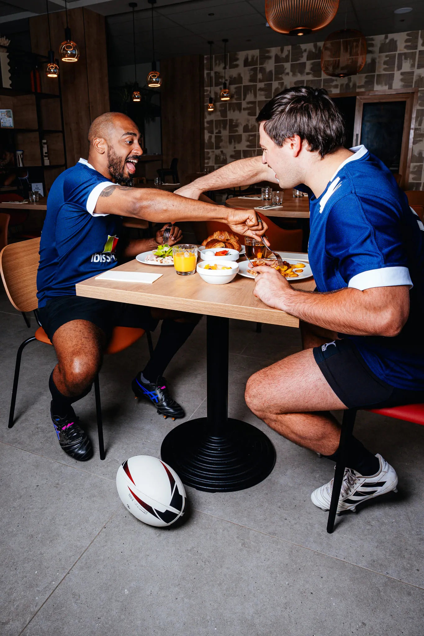 Deux joueurs de rugby en maillots bleus assis à une table dans un restaurant, riant et levant leur poing en signe de victoire, avec un ballon de rugby au sol près de leurs pieds.