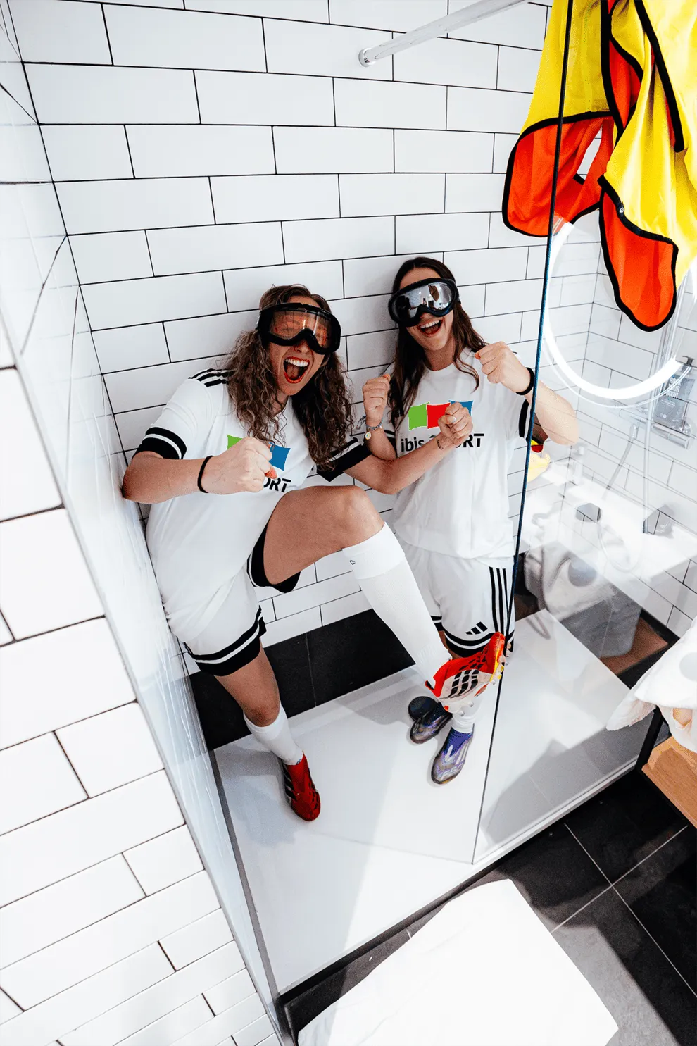 Deux personnes en tenue de sport blanche avec des lunettes de protection sont dans une douche à carreaux blancs, souriantes et faisant un geste de victoire, l'une levant une jambe avec des chaussures colorées.