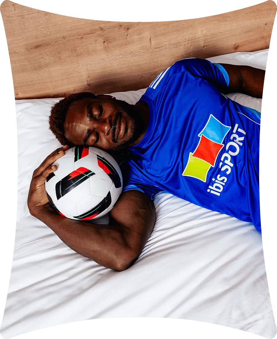Un homme noir portant un maillot bleu ibis sport dort sur un lit blanc en tenant un ballon de football sous sa tête.