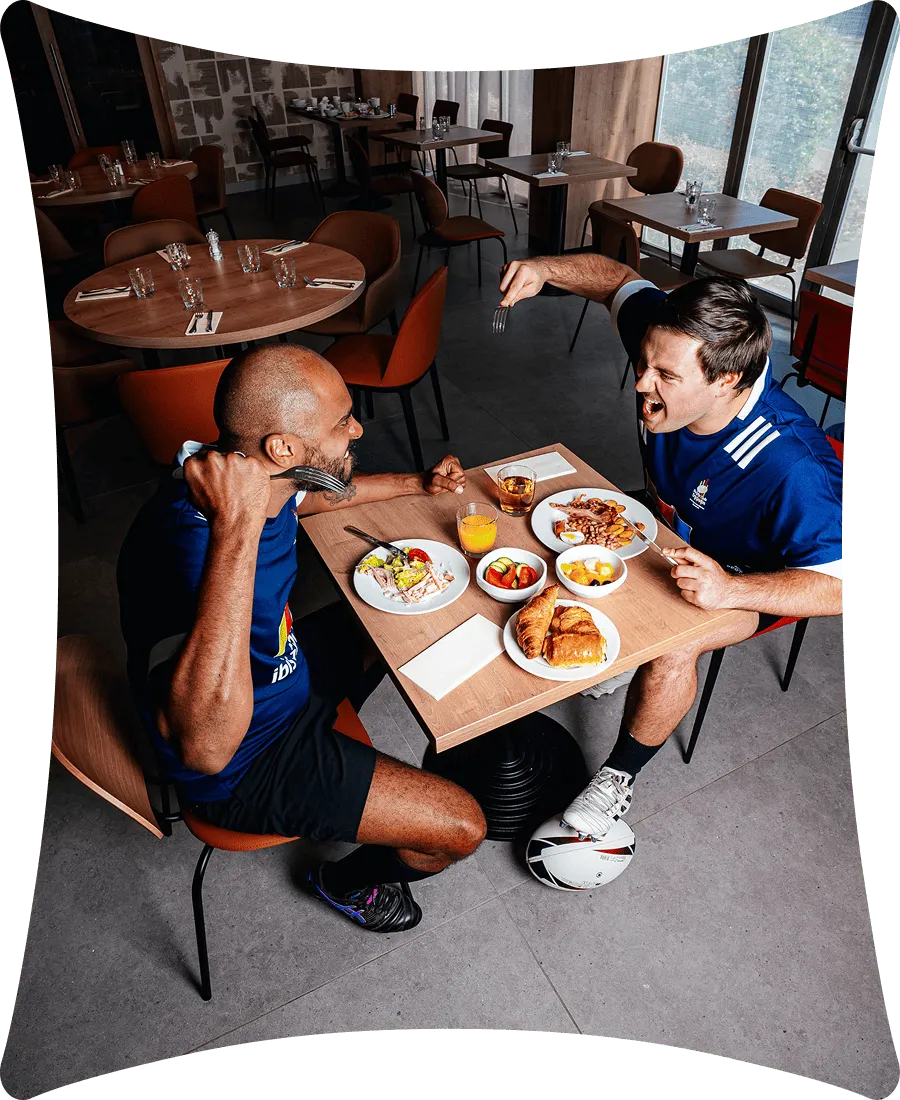Deux hommes en tenue de sport bleu sont assis à une table avec un petit-déjeuner comprenant croissants, fruits, jus d'orange et petit déjeuner anglais, dans une salle à manger vide.