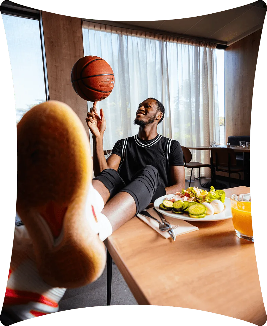 Un homme souriant en maillot noir fait tourner un ballon de basket sur le doigt tout en ayant les pieds sur une table avec une assiette de salade et un verre de jus d'orange à côté.