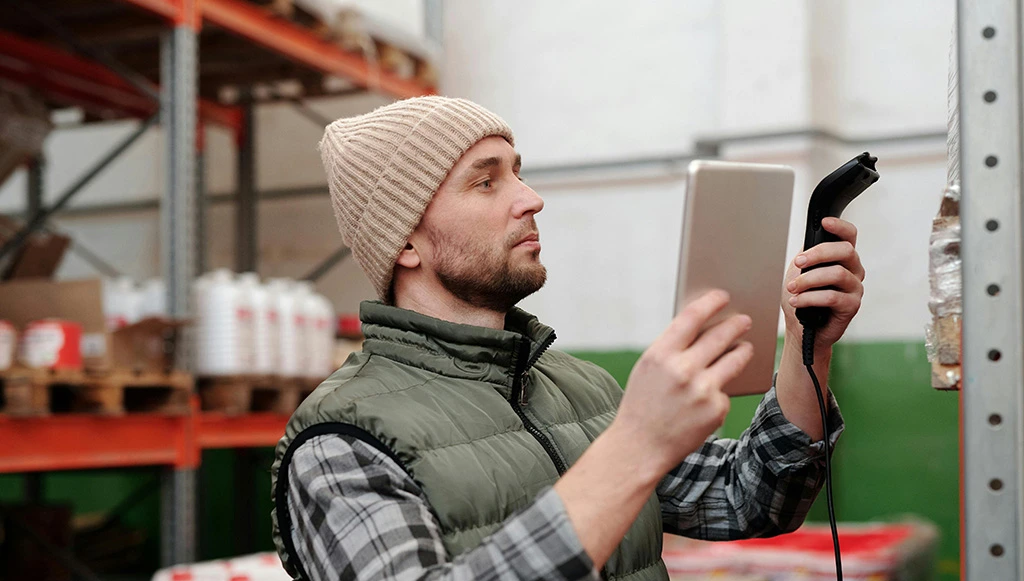 A warehouse worker wearing a beanie and padded vest scans a product box using a handheld barcode scanner and tablet in a storage facility.