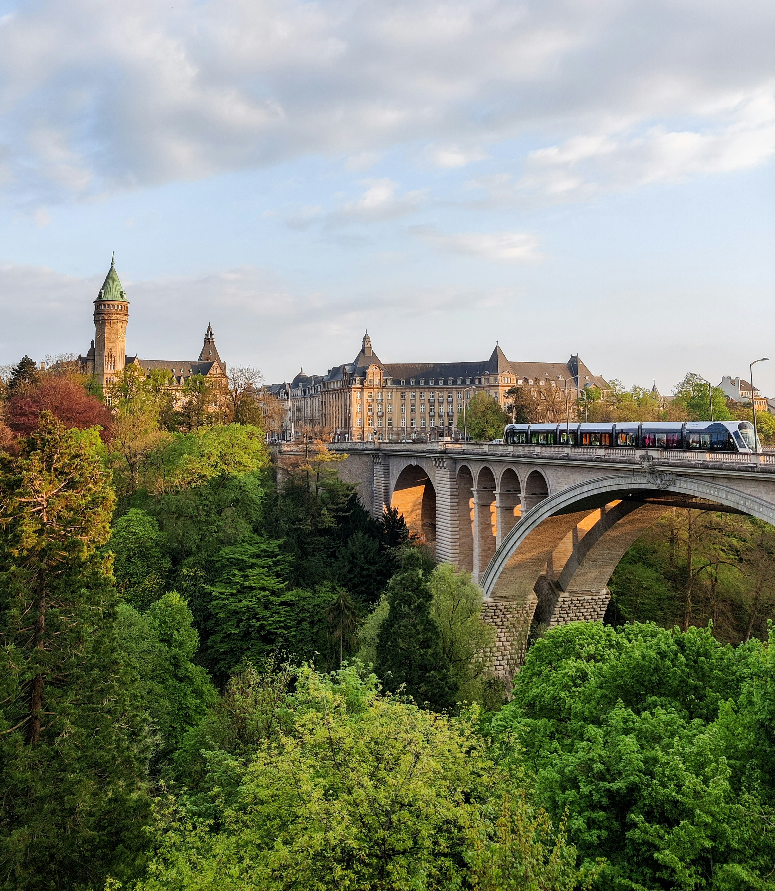 The image shows the Adolphe Bridge (Pont Adolphe) crossing the Pétrusse River Valley in Luxembourg City, with a modern tram passing over it. 