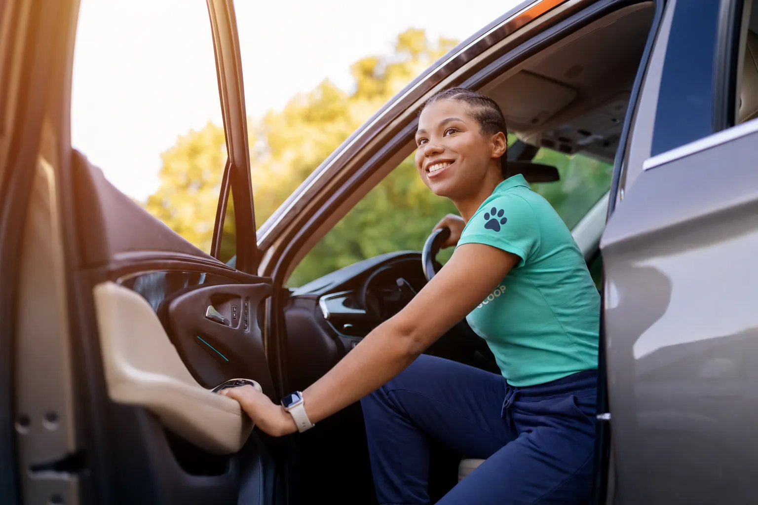Smiling woman wearing a teal shirt with a paw print on the sleeve, seated in a car with the door open, holding the steering wheel and looking outside.