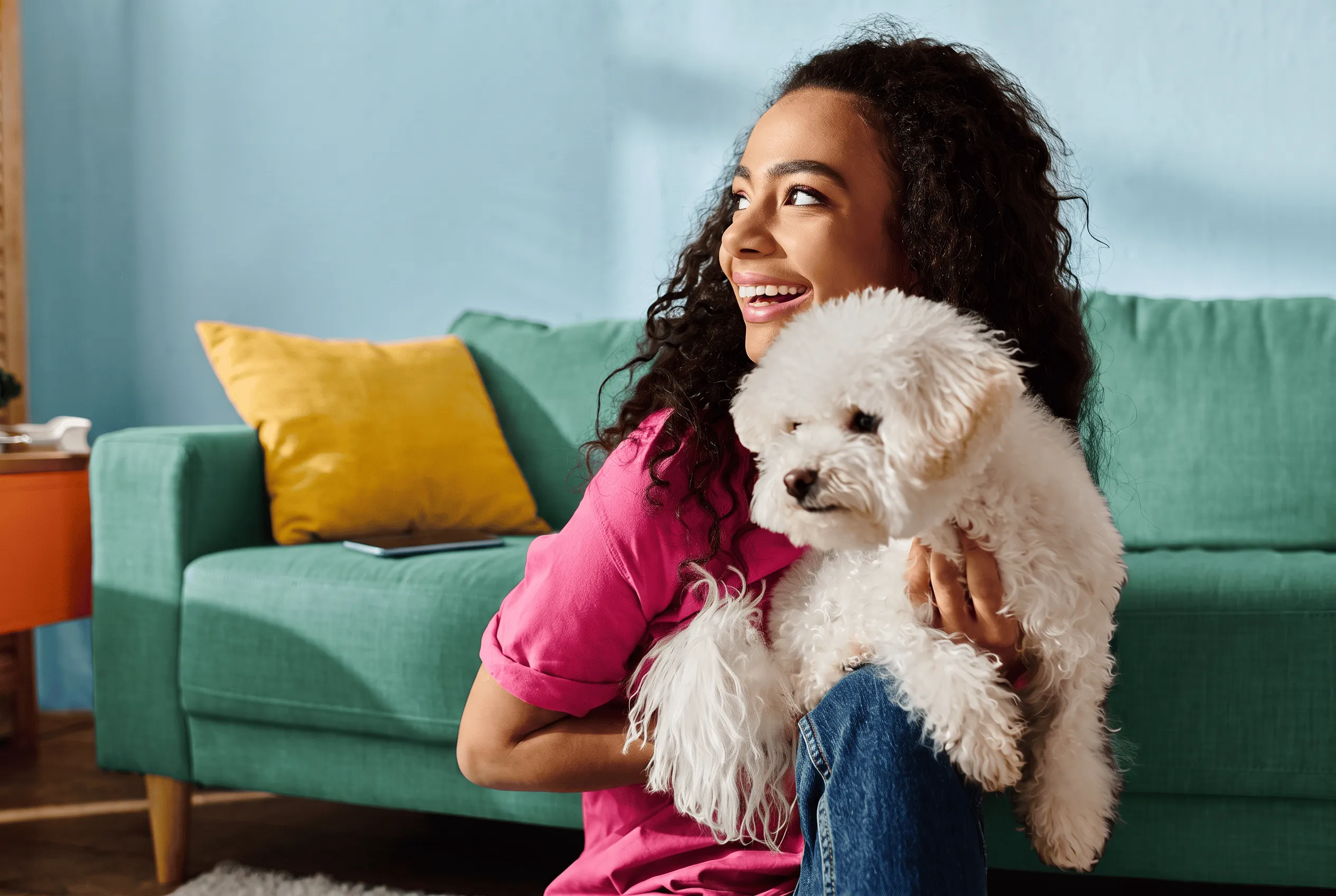 Smiling woman in pink shirt sitting on the floor indoors, holding a small fluffy white dog, with a green couch and yellow pillow in the background.