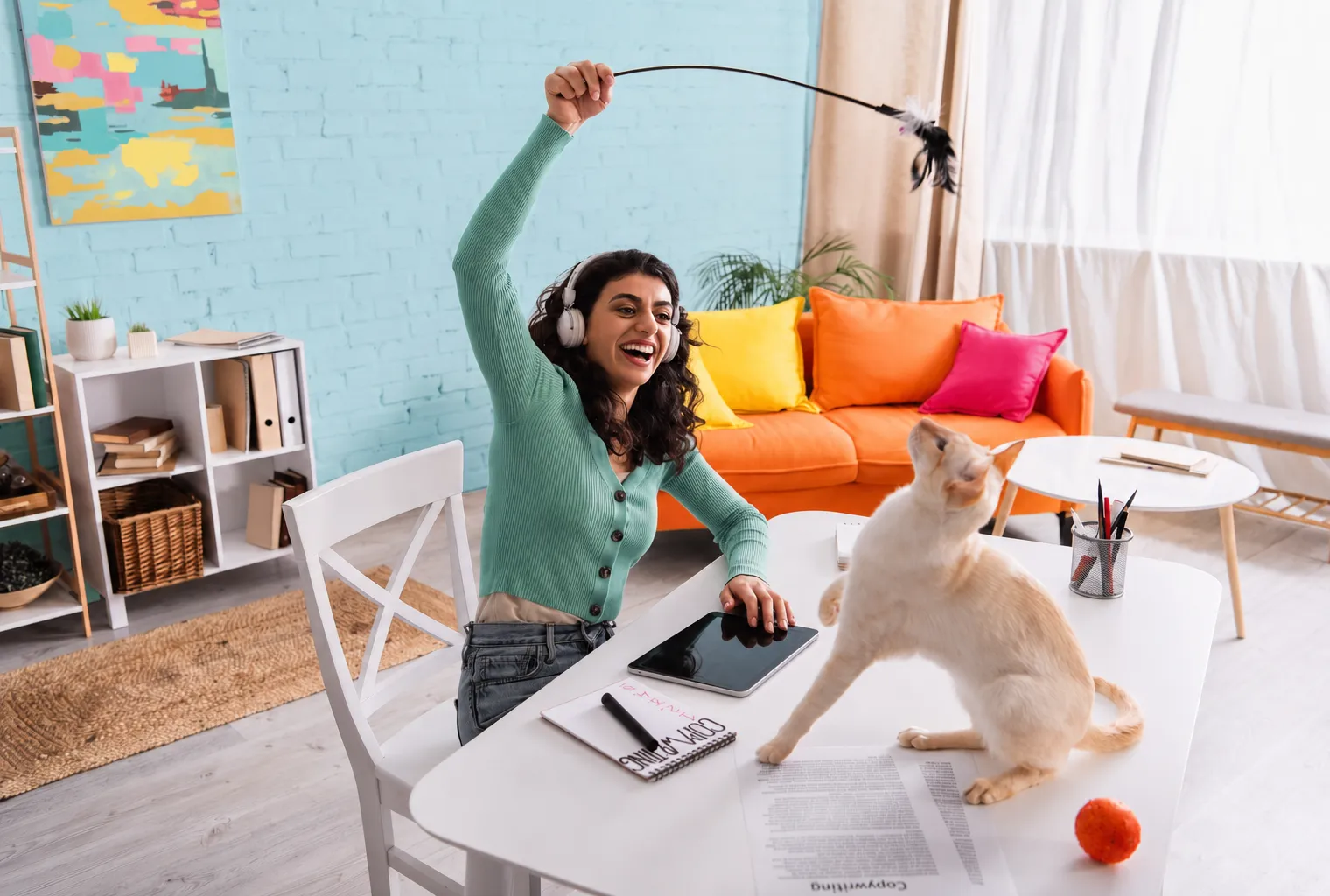 Woman wearing headphones and a green sweater playing with a cream-colored cat using a feather toy while sitting at a white table in a colorful living room with an orange couch and bright pillows.