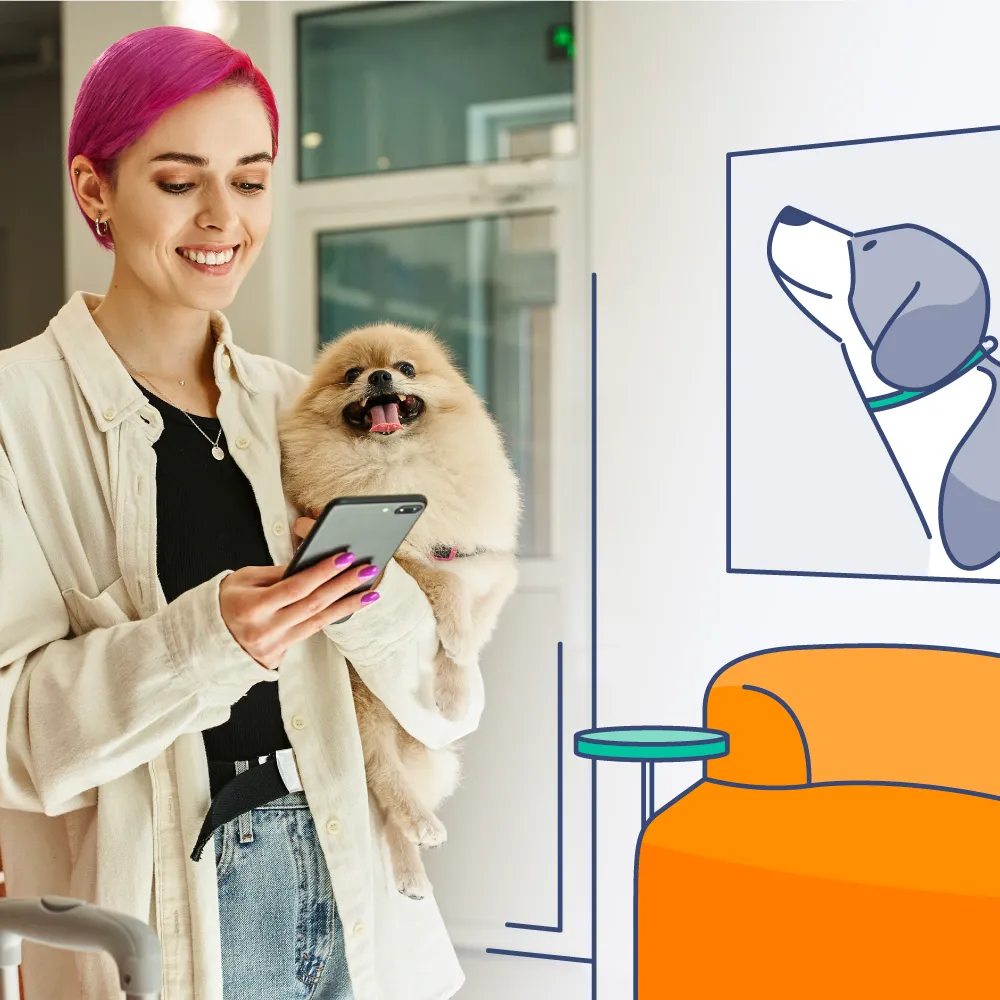 Smiling woman with bright pink hair holding a small fluffy dog while looking at a smartphone, standing indoors near a wall with a cartoon dog illustration and an orange chair.