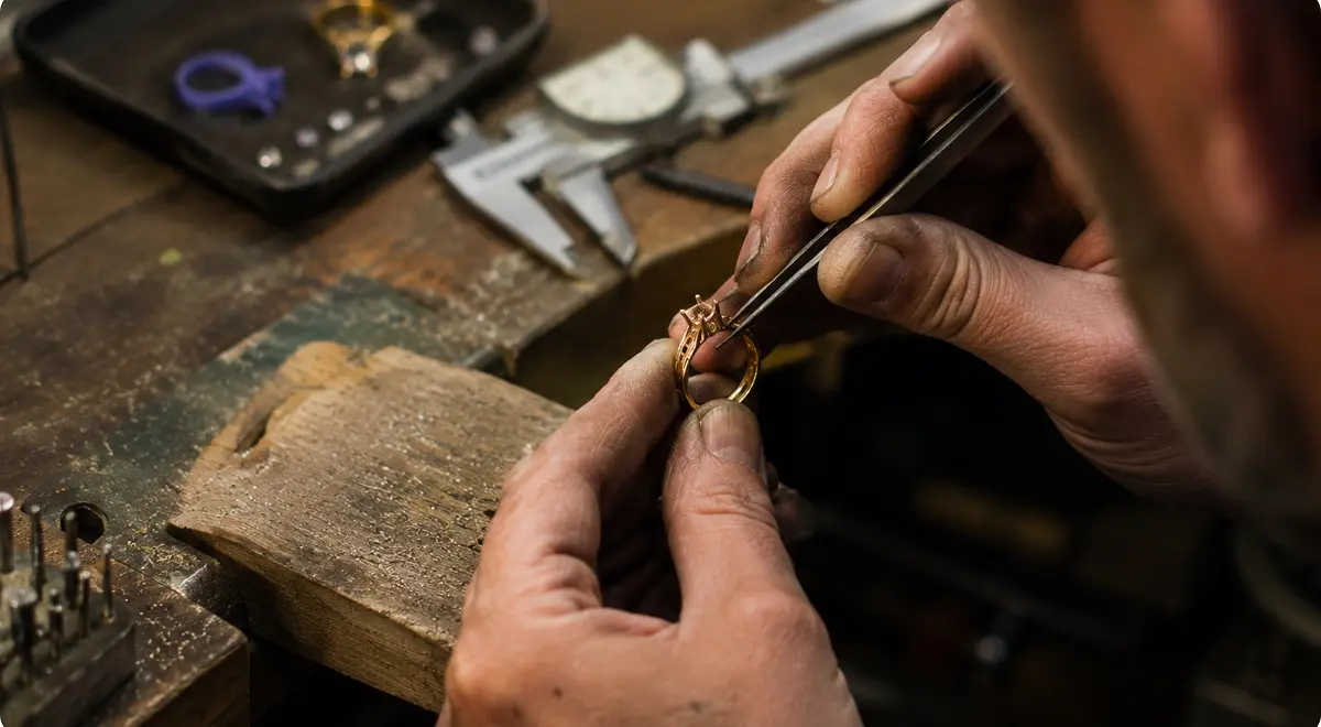 Joaillier sertissant une pierre dans une bague en or à la brucelle sur son établi, pied à coulisse et limes à portée de main.