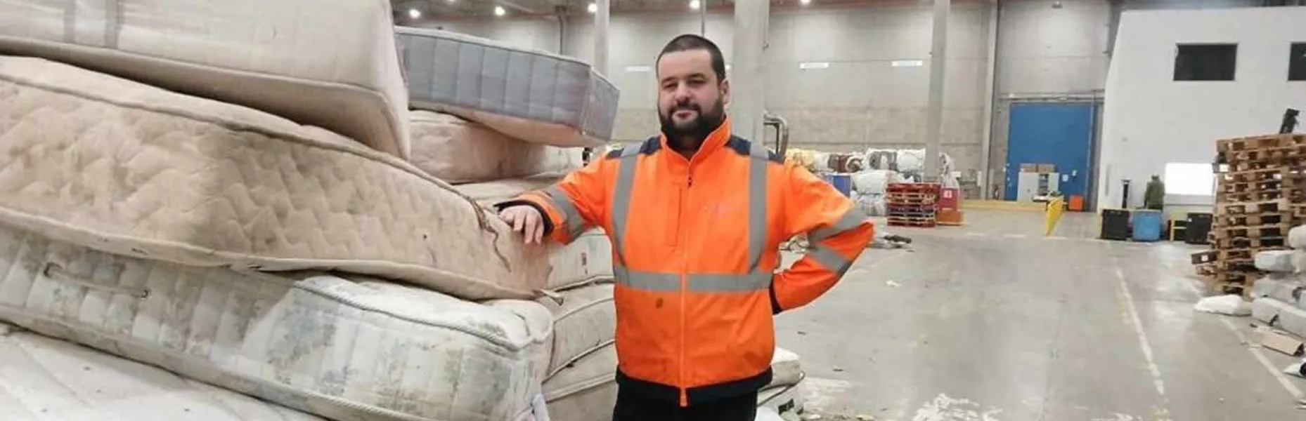 A man in a hi vis jacket standing beside a pile of used mattresses in a warehouse.