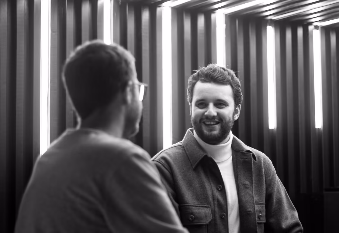 Two men engaged in conversation in a modern setting with vertical light strips on the walls, one man facing forward smiling and the other seen from behind.