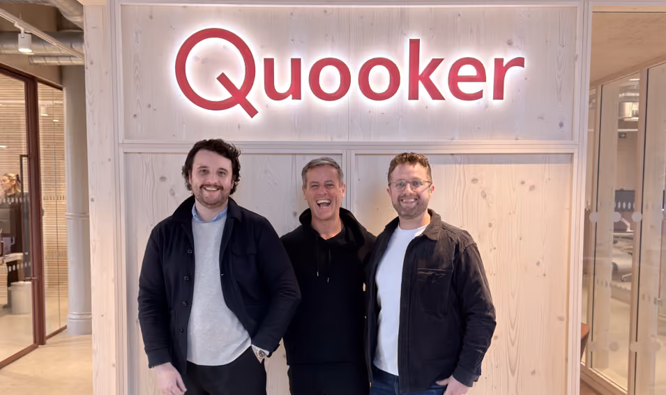 Three men standing indoors smiling in front of a light wooden wall with the illuminated Quooker logo above them.