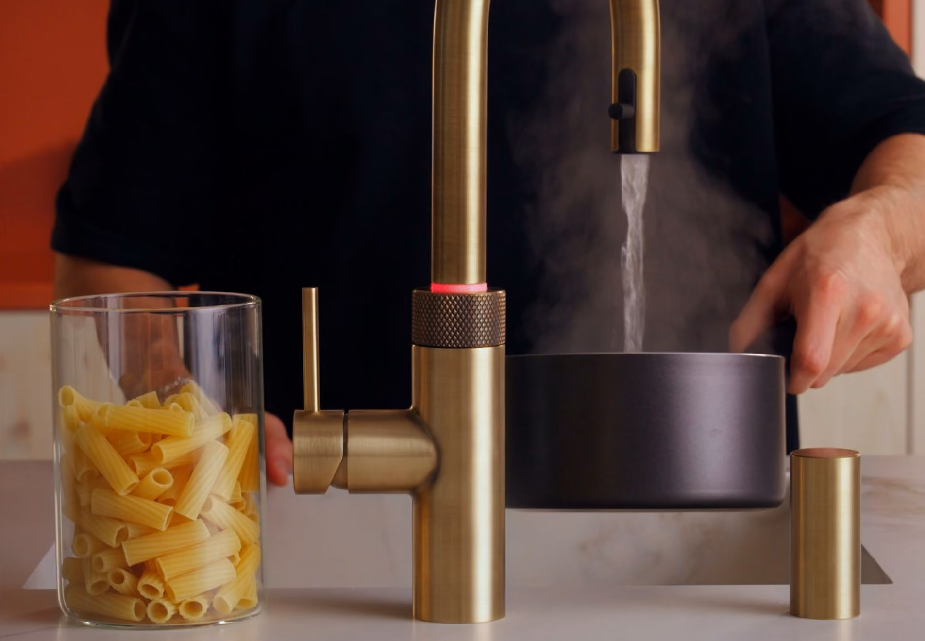 Brass kitchen faucet running hot water with steam rising into a black pot held by person wearing black on a countertop next to a clear container filled with uncooked rigatoni pasta.