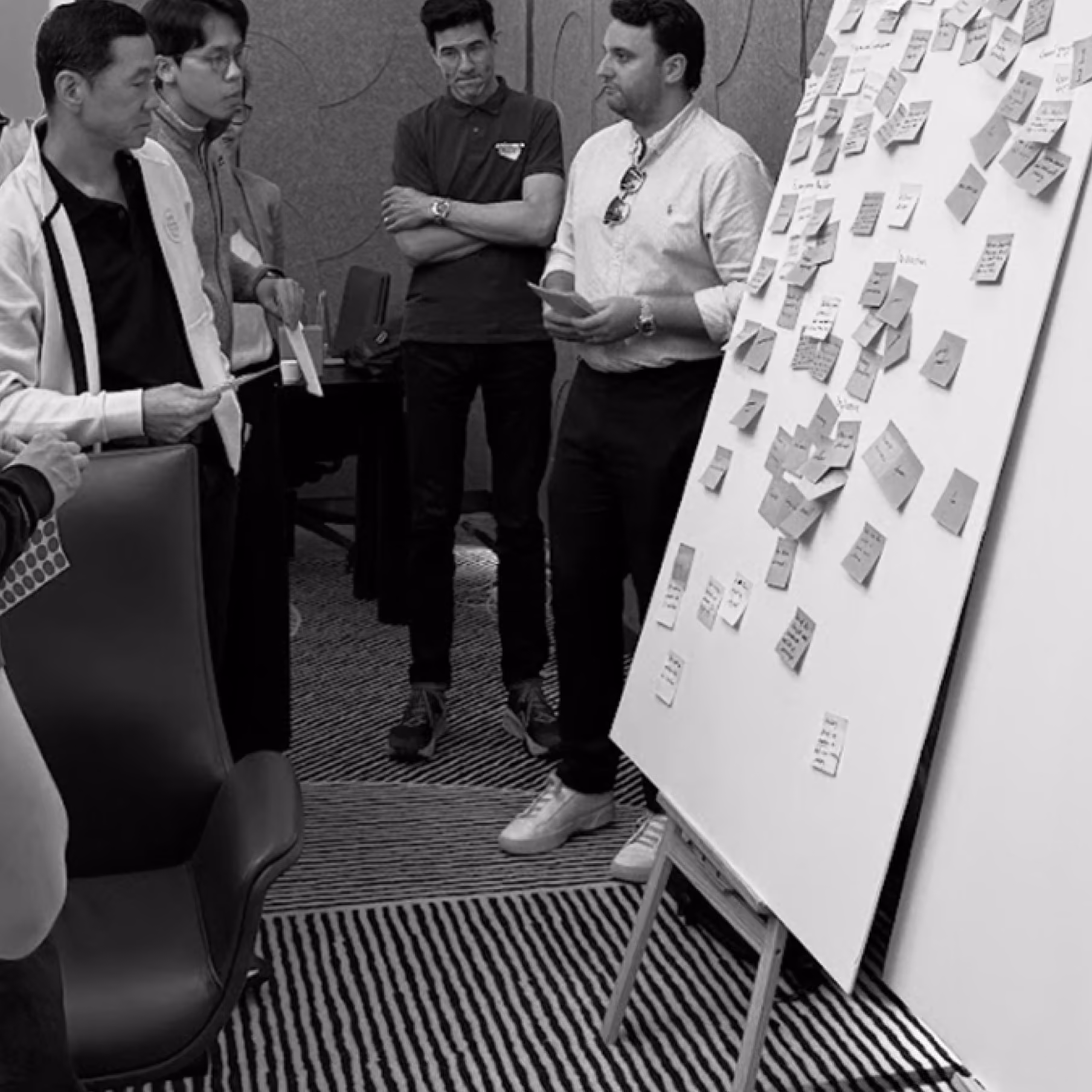 Four men stand around a whiteboard covered with numerous sticky notes in a modern office setting, engaging in a discussion or brainstorming session.